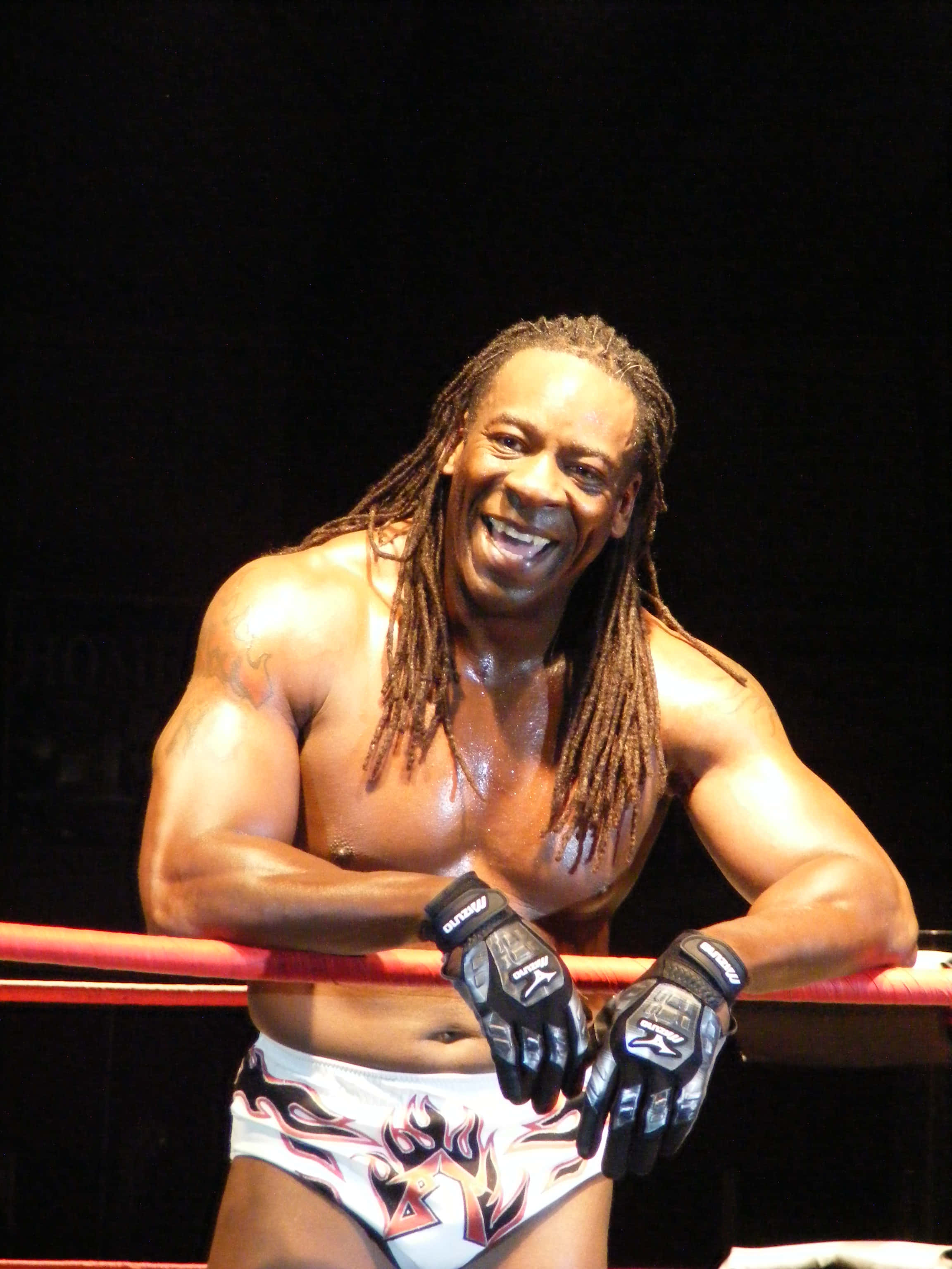 Booker T During TNA Live Event 壁紙 — 一位肌肉發達、留著長辮子的摔角手，在明亮的舞台燈光下靠著紅色擂台繩索咧嘴大笑。