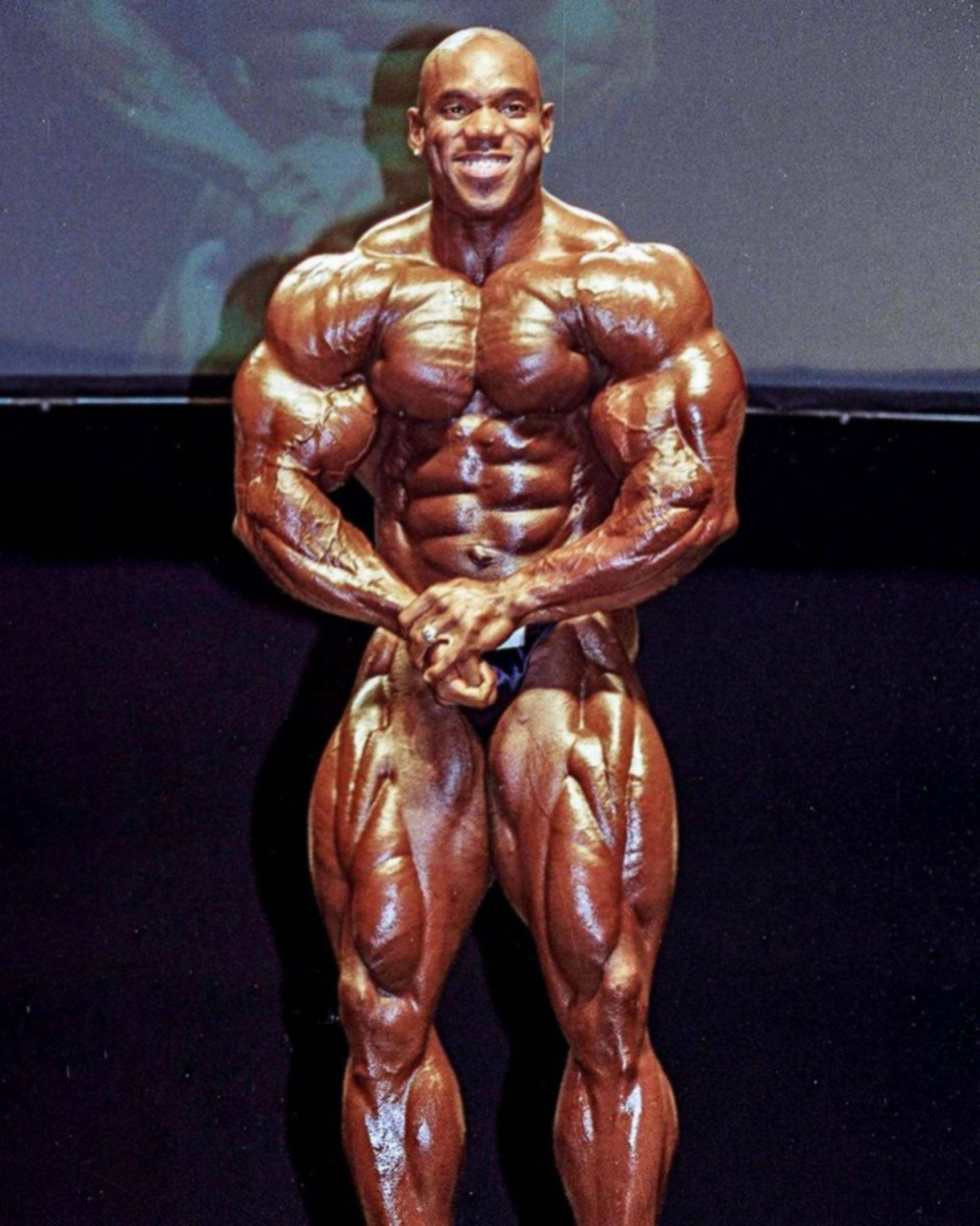 Masculine Flex Wheeler 壁紙 — 一位肌肉發達、皮膚塗油的健美運動員在黑暗舞台上自信地擺姿勢，燈光明亮。