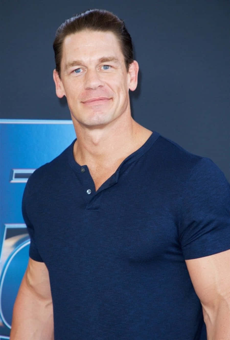 John Cena 桌布 壁紙 — 一位肌肉發達、藍眼睛、深色頭髮的男子，身穿海軍藍亨利衫，在深藍色背景前自信地微笑。