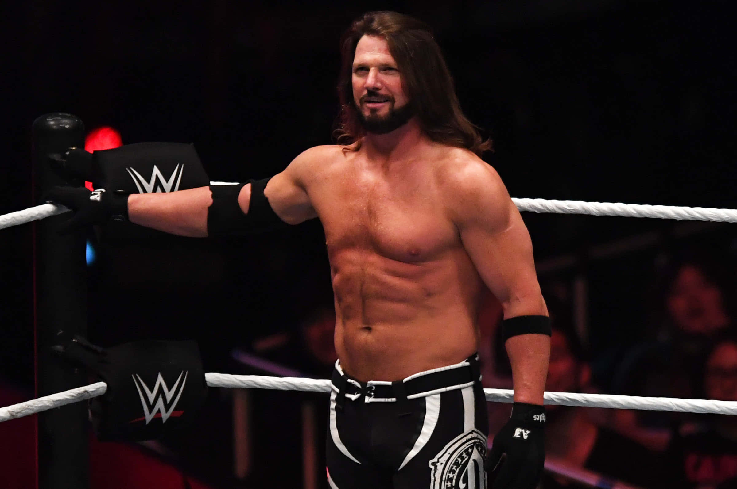 AJ Styles WWE Smackdown 壁紙 — 一位肌肉發達、長髮鬍鬚的摔角手自信地站在摔角場上，穿著黑白短褲和手套。