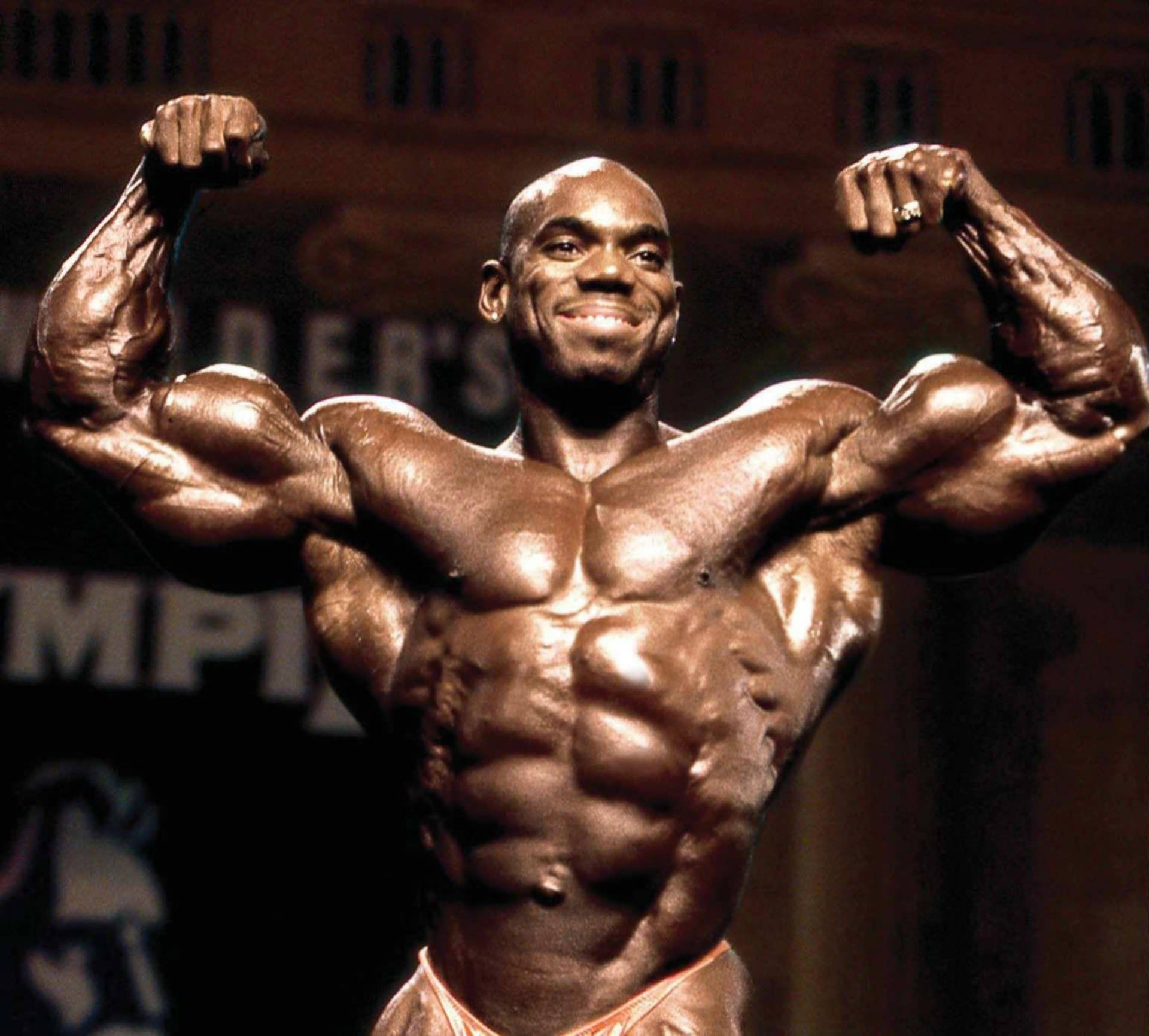 Husky Flex Wheeler 壁紙 — 一位肌肉發達的健美運動員在舞台燈光下自信微笑，繃緊手臂和胸部。