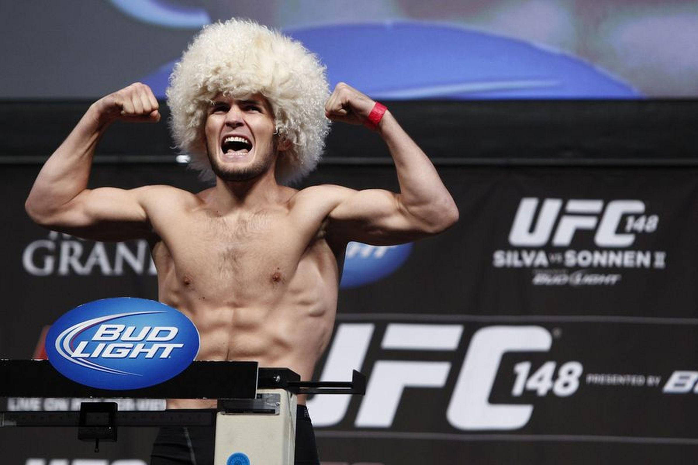 UFC 桌布 壁紙 — 一位肌肉發達的戰士戴著白色捲曲假髮，在UFC稱重台上彎曲手臂並咆哮。