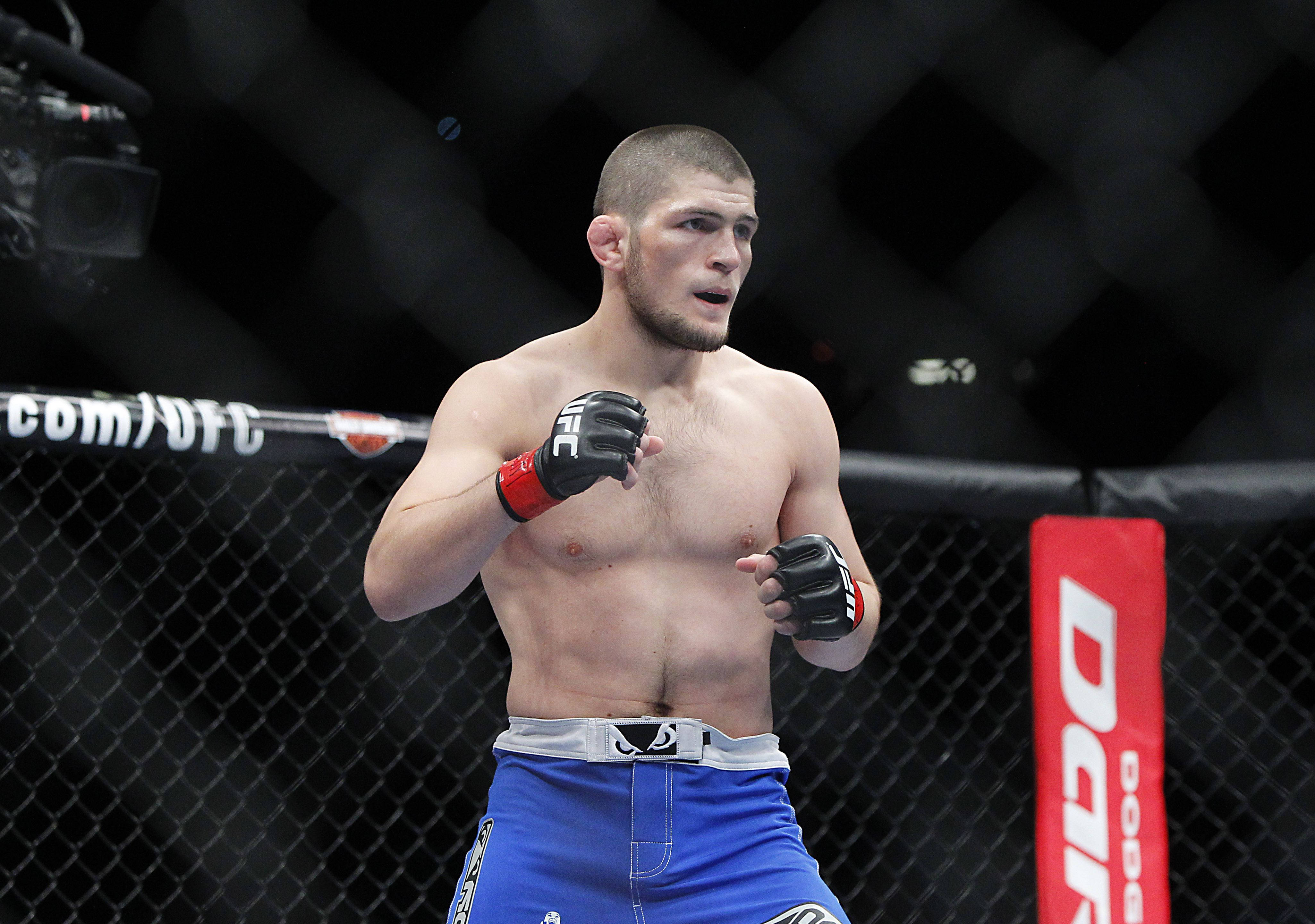UFC 桌布 壁紙 — 一位肌肉發達的戰士穿著藍色短褲和黑色手套，在UFC八角籠內警覺地站立，鐵絲網圍欄和攝像機清晰可見。