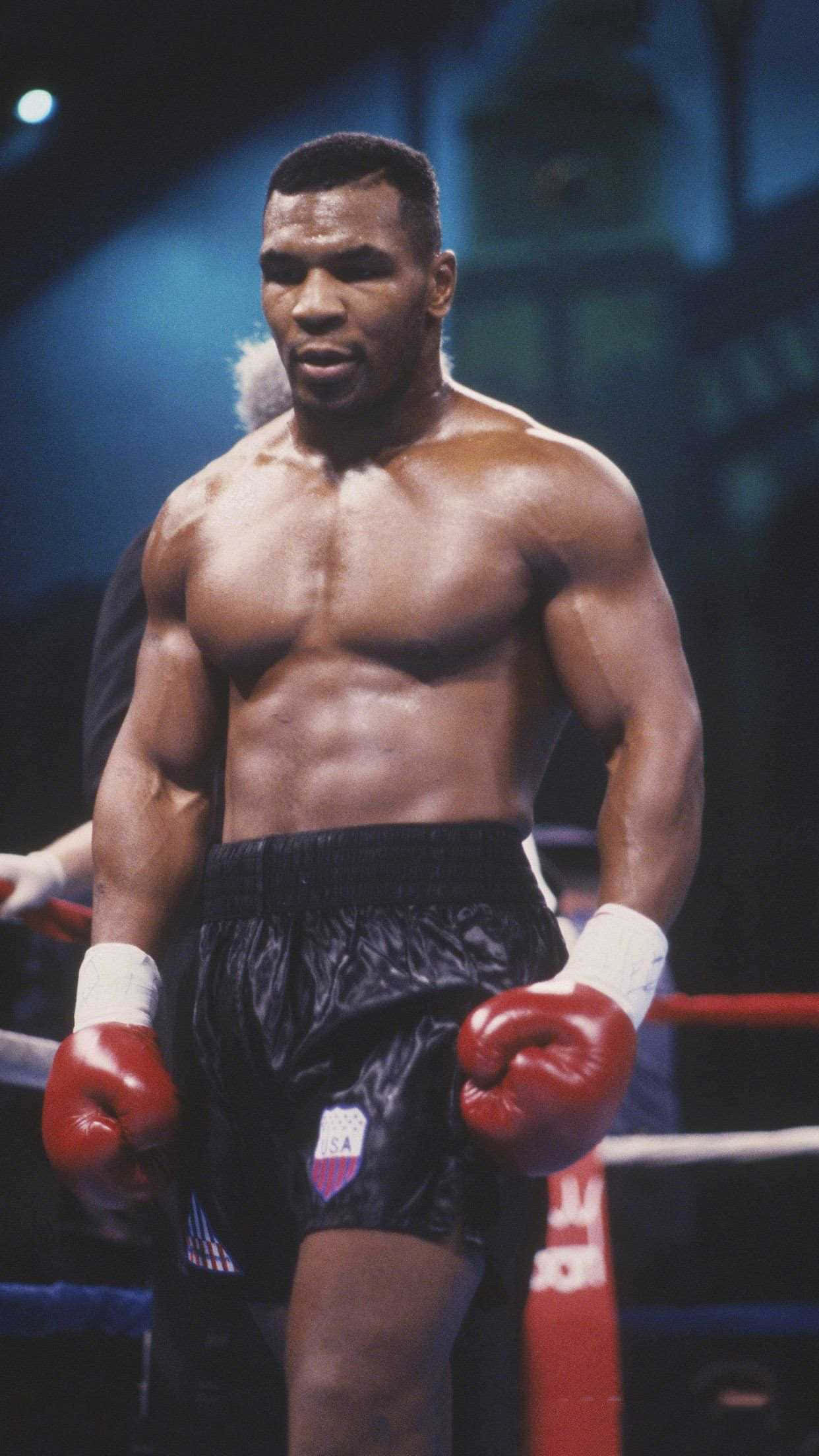 mike tyson 桌布 壁紙 — 一位肌肉發達的拳擊手戴著紅色手套和黑色短褲，在燈光照射的擂台上，背景有繩索和模糊的人群，神情專注。