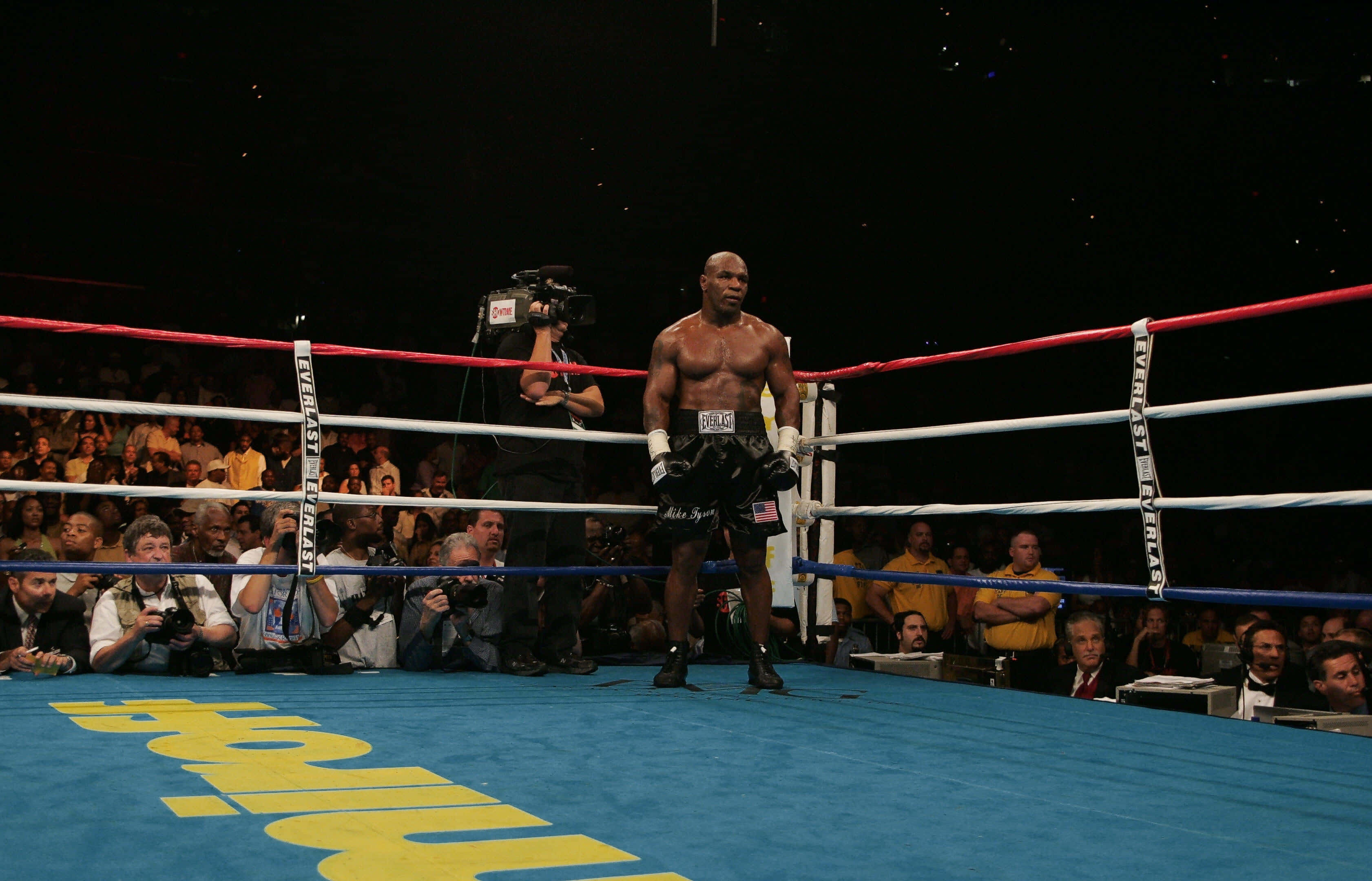 mike tyson 桌布 壁紙 — 一位肌肉發達的拳擊手獨自站在燈光明亮的擂台上，攝影師和觀眾在場邊專注地觀看。