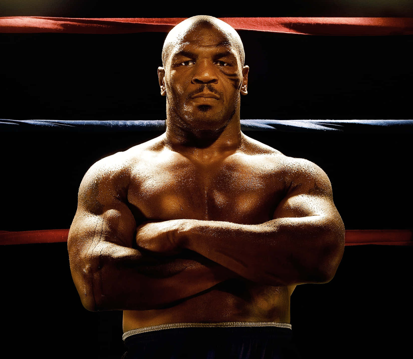 mike tyson 桌布 壁紙 — 一位肌肉發達的拳擊手自信地站在燈光昏暗的擂台上，雙臂交叉，汗水在戲劇性的燈光下閃閃發光。