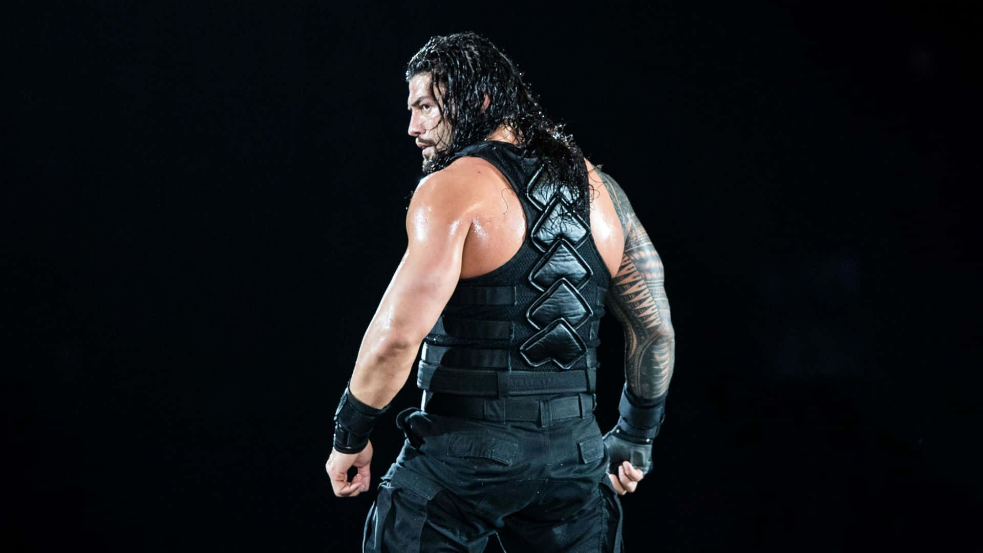 Roman Reigns 桌布 壁紙 — 一位肌肉發達的摔角手，濕髮和紋身，穿著黑色背心，在黑暗背景前回頭看。