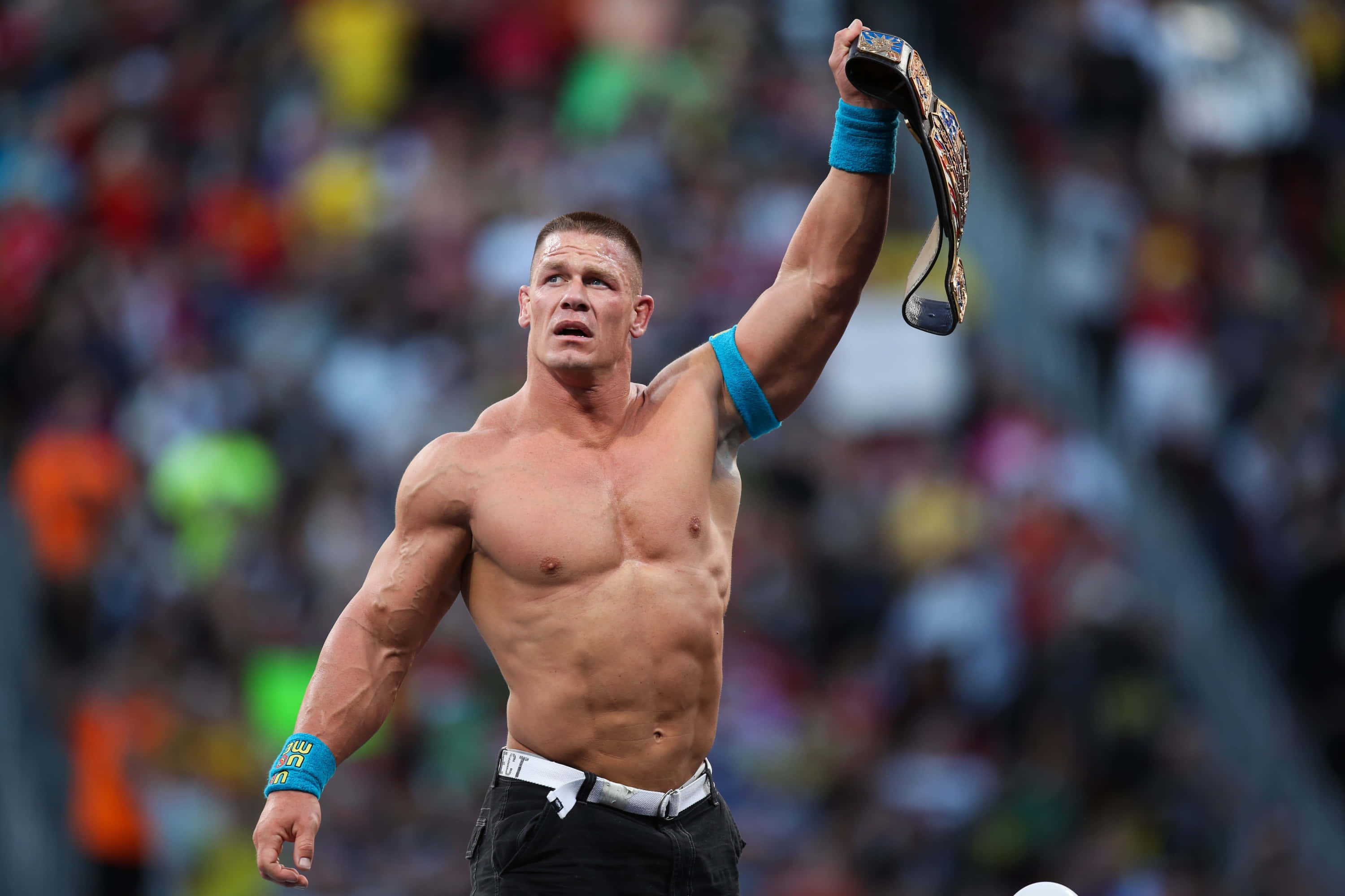 John Cena 桌布 壁紙 — 一位肌肉發達的摔角手在模糊的歡呼人群面前，勝利地將冠軍腰帶高舉過頭頂。