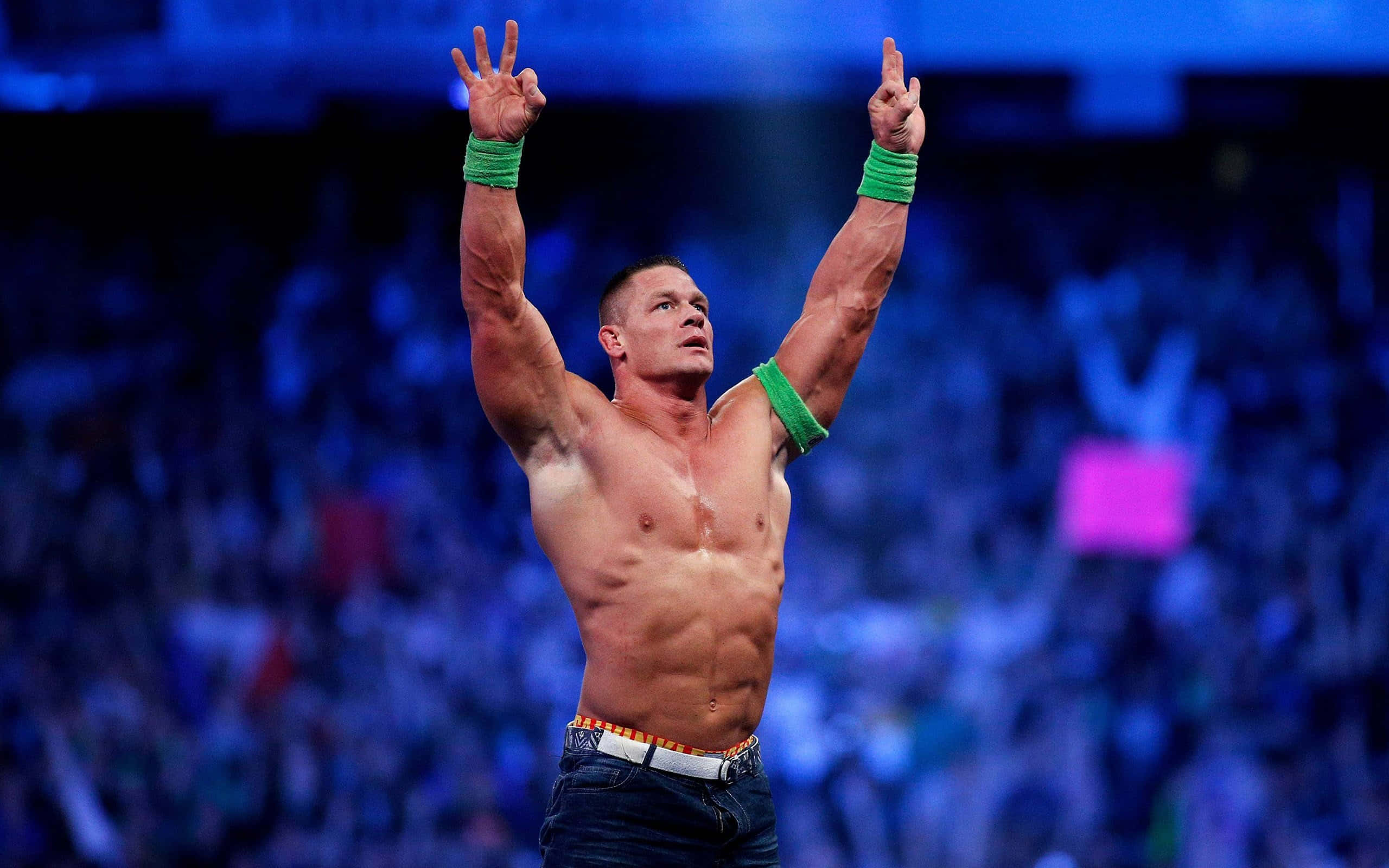 WWE 桌布 壁紙 — 一位肌肉發達的摔角手戴着綠色護腕，在藍色場館燈光下，於模糊的歡呼人群前高舉雙臂，展現勝利姿態。