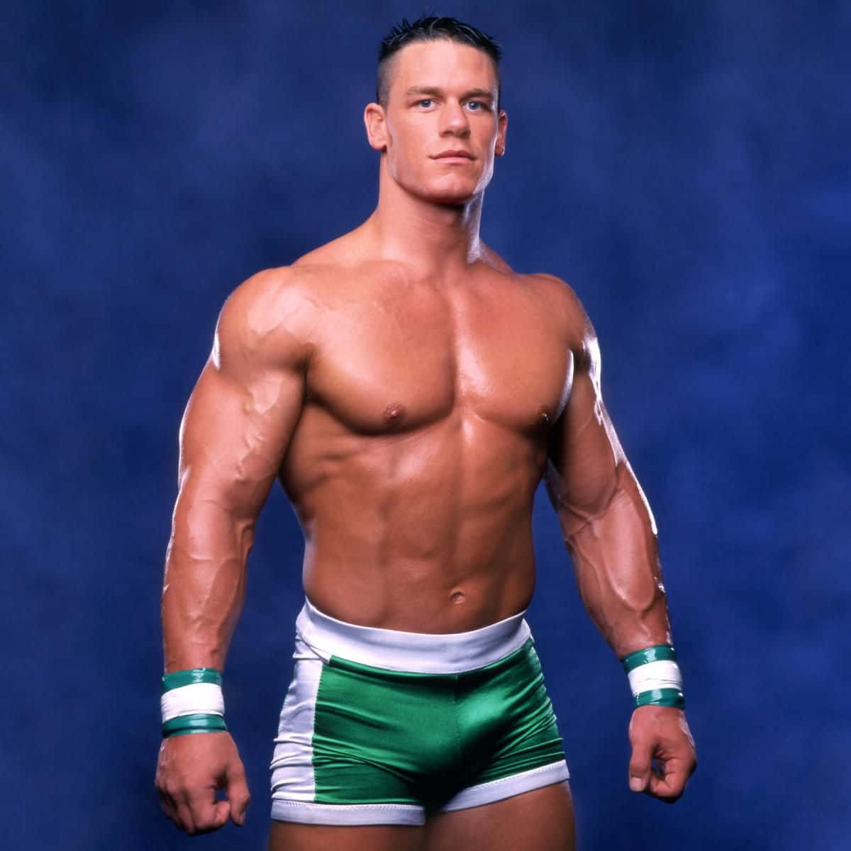John Cena 桌布 壁紙 — 一位肌肉發達的摔角手穿著綠色和白色的短褲，在煙霧繚繞的藍色背景下自信地站立，體格分明，眼神凌厲。