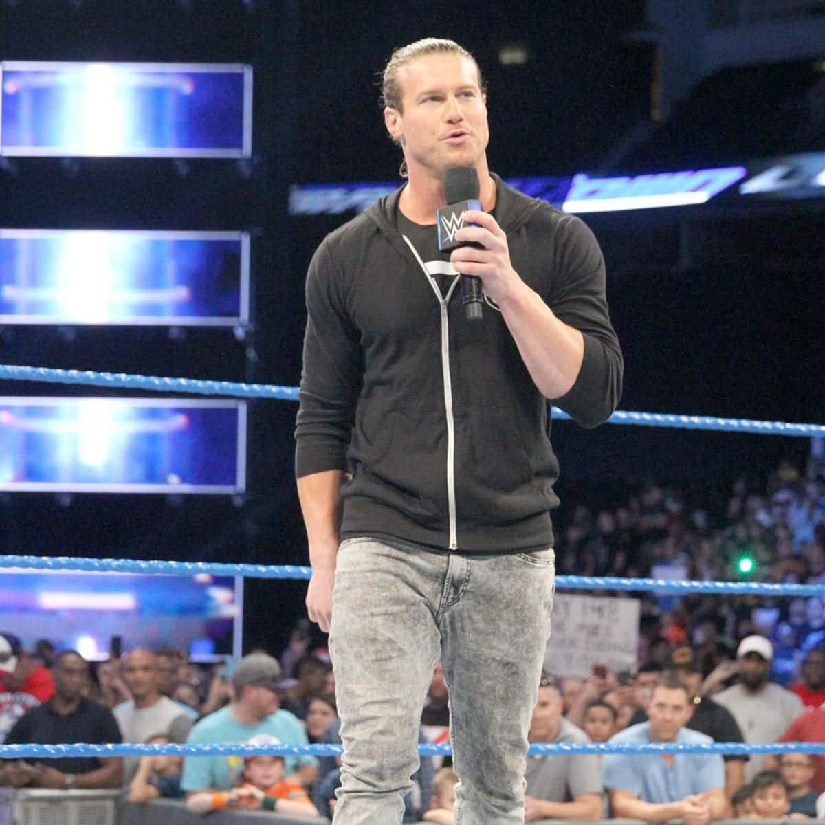 Dolph Ziggler WWE Wrestler 壁紙 — 一位肌肉發達的摔角手穿著黑色連帽衫，站在摔角場上，手持麥克風，在明亮的場館燈光下向歡呼的人群講話。