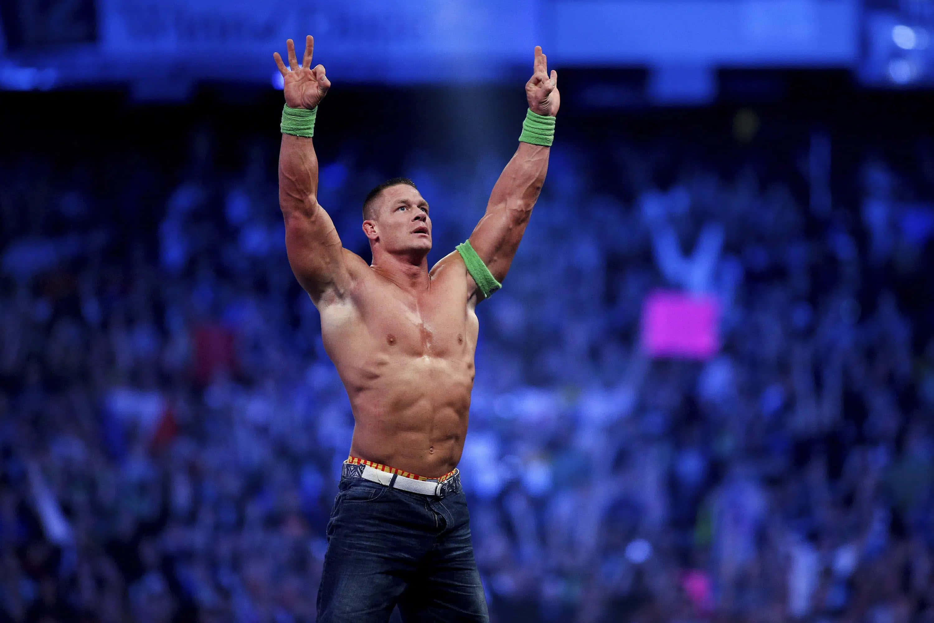 John Cena 桌布 壁紙 — 一位肌肉發達的摔角手身穿牛仔褲和綠色護腕，在藍色燈光下擠滿人的場館中高舉雙臂慶祝勝利。