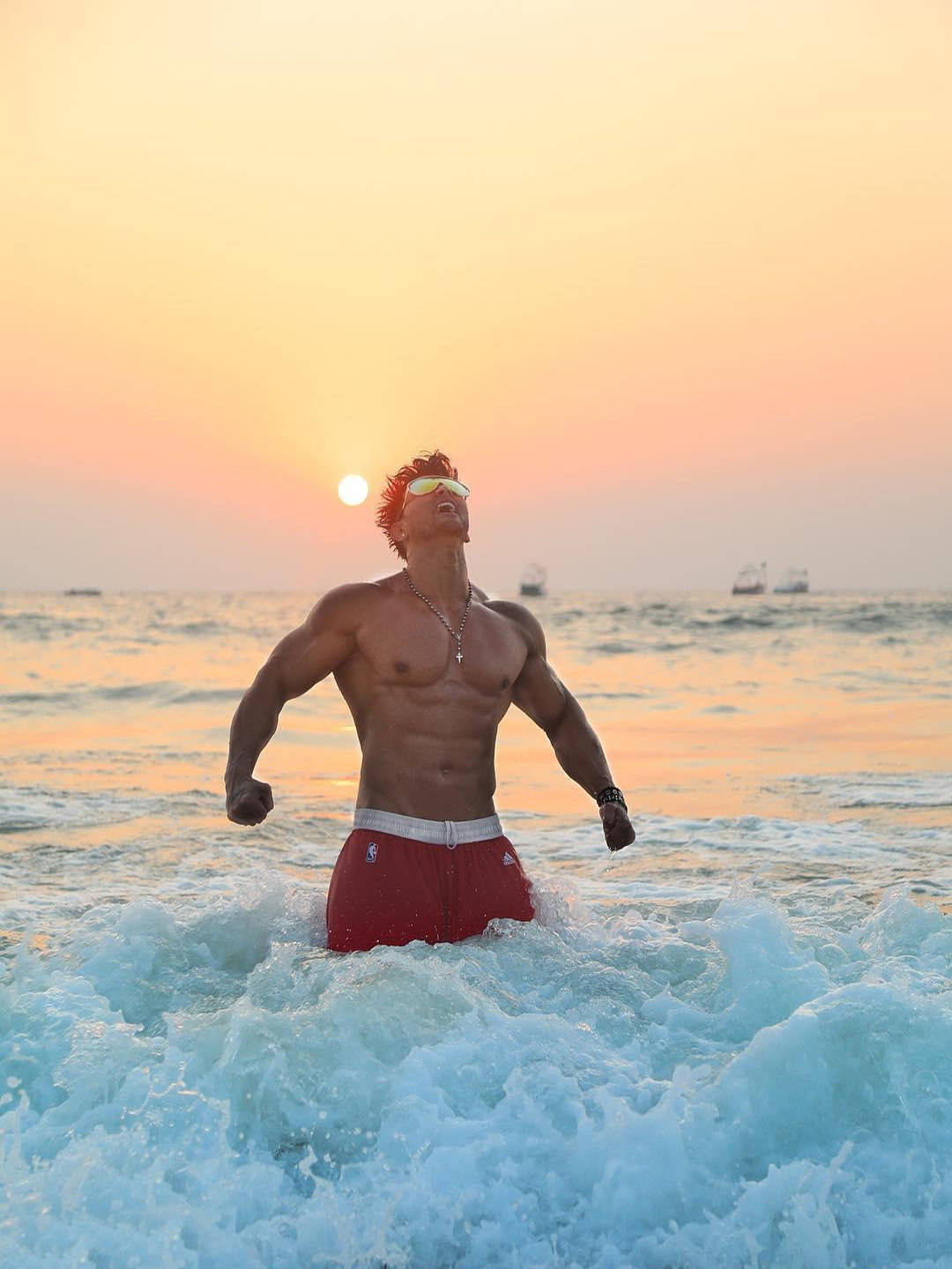 India 桌布 壁紙 — 一位肌肉發達的男子穿著紅色泳褲，在夕陽下站在海浪中，遠處有船隻，姿態勝利。