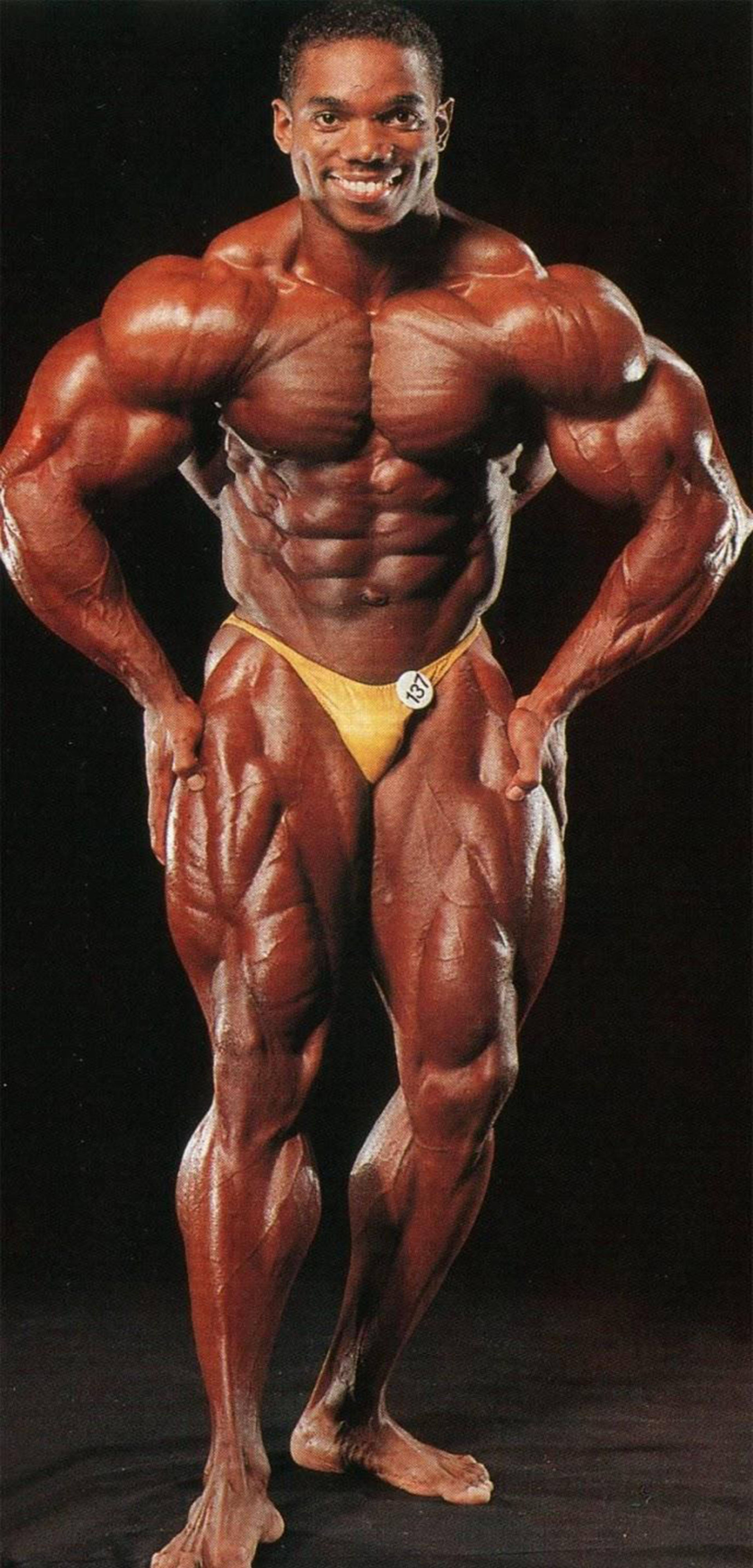 Beefy Flex Wheeler 壁紙 — 一位肌肉發達的男性健美運動員自信地穿著黃色比賽褲在黑色背景前擺姿勢。