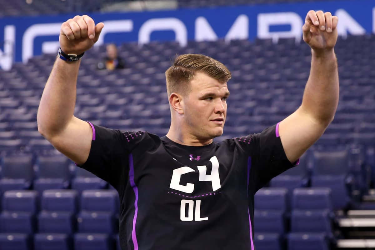 Mike Mcglinchey 桌布 壁紙 — 一位肌肉發達的足球員身穿黑色和紫色球衣，在空無一人的體育館中勝利地高舉雙拳。