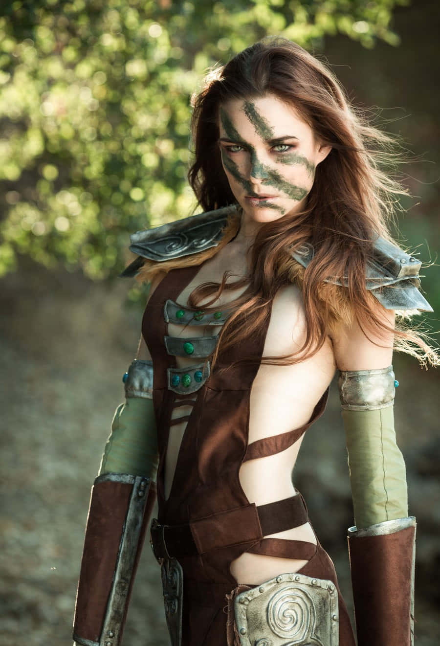 Aela The Huntress 桌布 壁紙 — 一位臉上塗有綠色顏料、身穿棕色鎧甲的女戰士自信地站在陽光明媚的森林中。