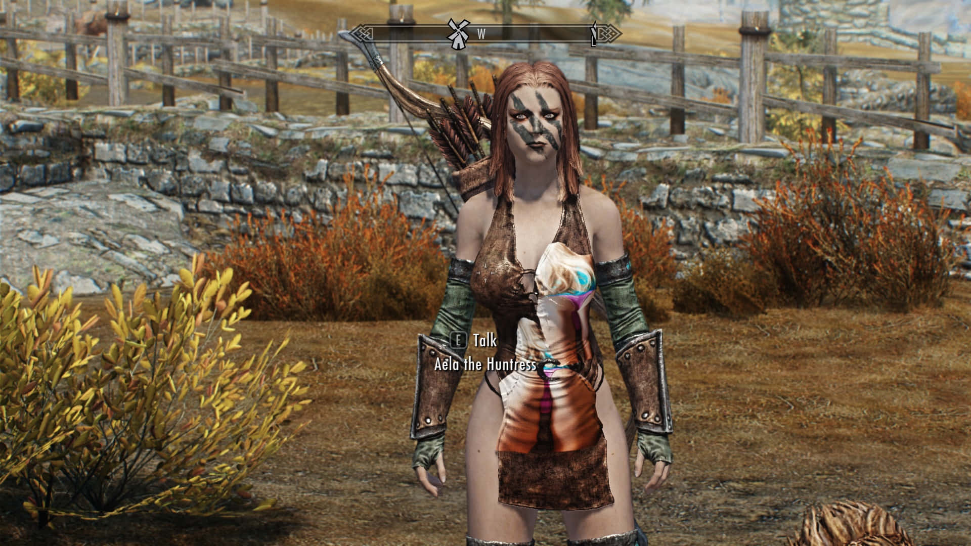 Aela The Huntress 桌布 壁紙 — 一位臉上塗有顏料、手持弓箭的女獵人，站在一個有秋日灌木叢的鄉村石柵欄戶外區域。