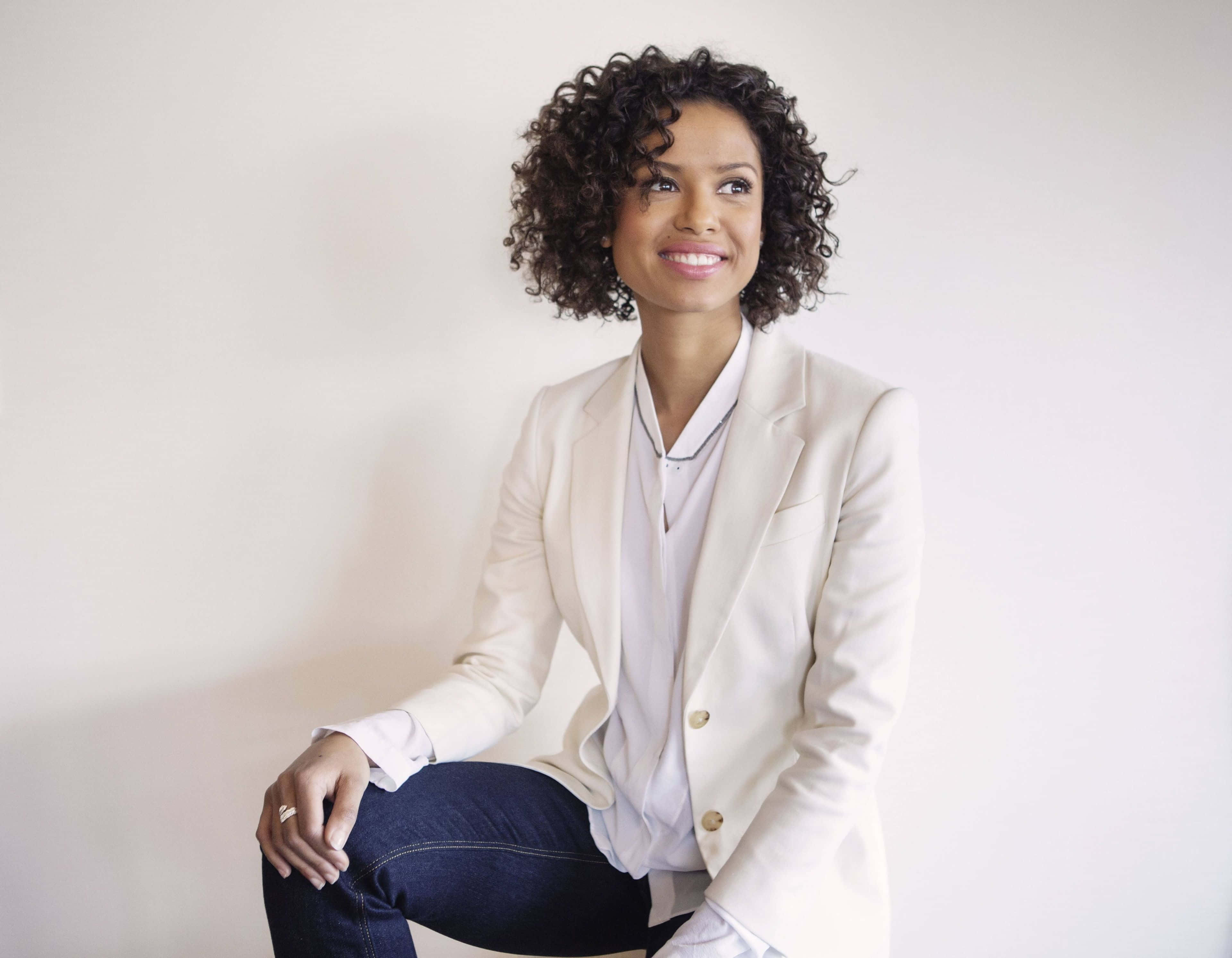 Gugu Mbatha-raw 桌布 壁紙 — 一位自信嘅捲髮女士，身穿米色西裝外套，坐喺純白牆前面微笑。