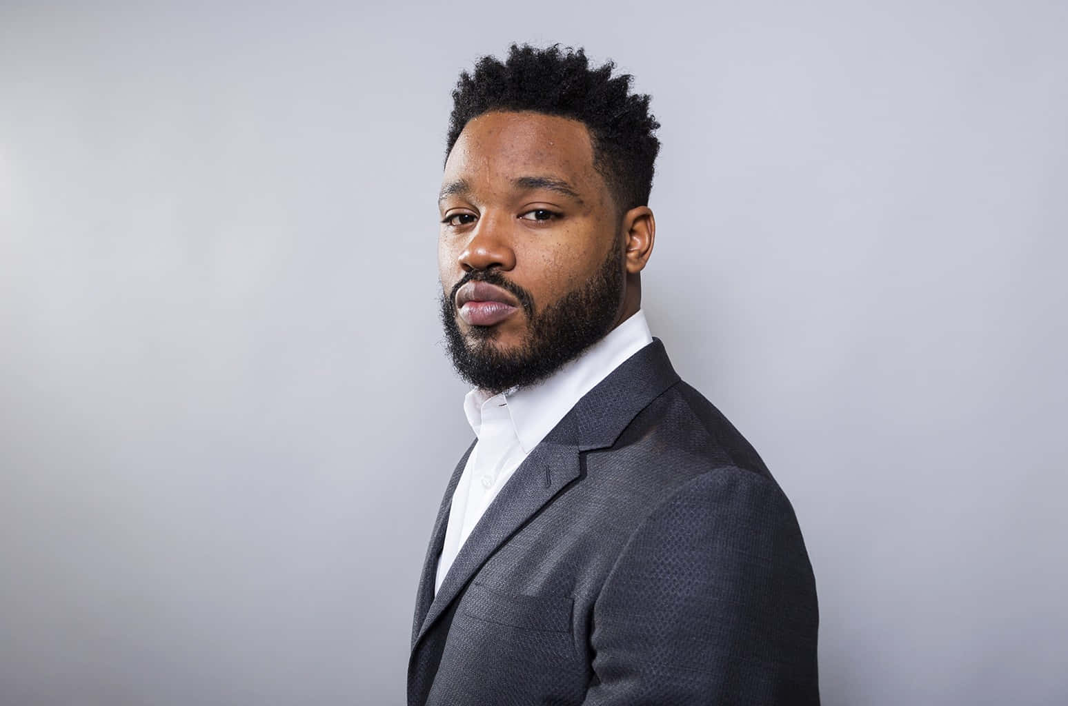 Ryan Coogler 桌布 壁紙 — 一位自信嘅男人，留鬍鬚，著住有質感嘅西裝，企喺純灰色背景前面，係專業攝影棚肖像。