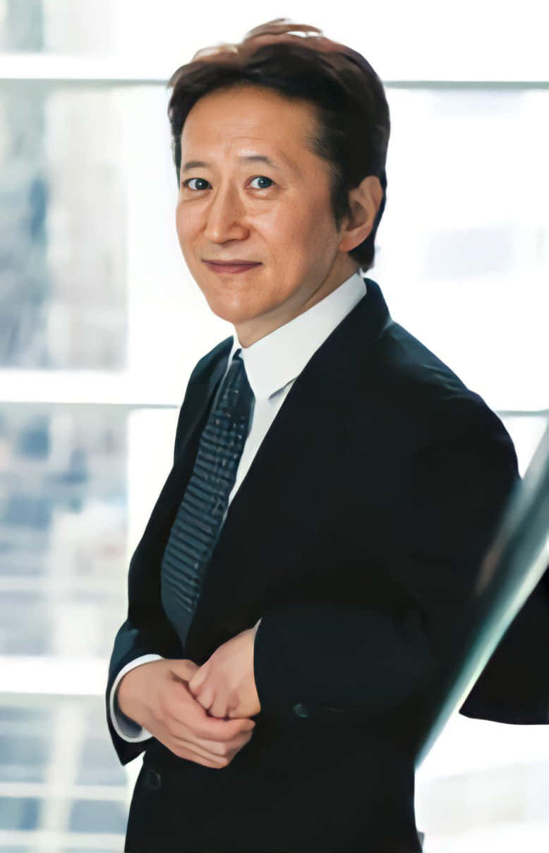 Hirohiko Araki 桌布 壁紙 — 一位自信的亞洲商人穿著俐落的黑色西裝，雙手交握，在明亮的窗戶背景下溫暖地微笑。