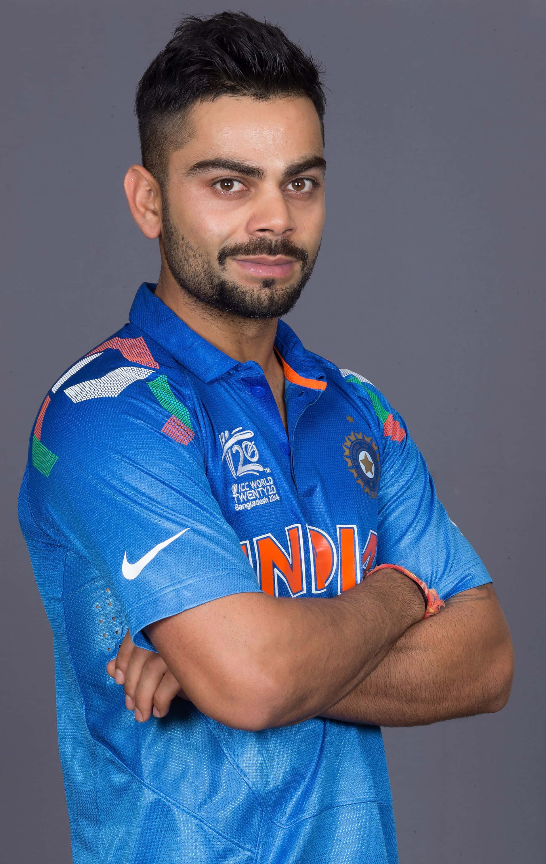 Virat Kohli 桌布 壁紙 — 一位自信的印度板球隊隊長身穿藍色隊服，雙臂交叉，站在純灰色背景前。