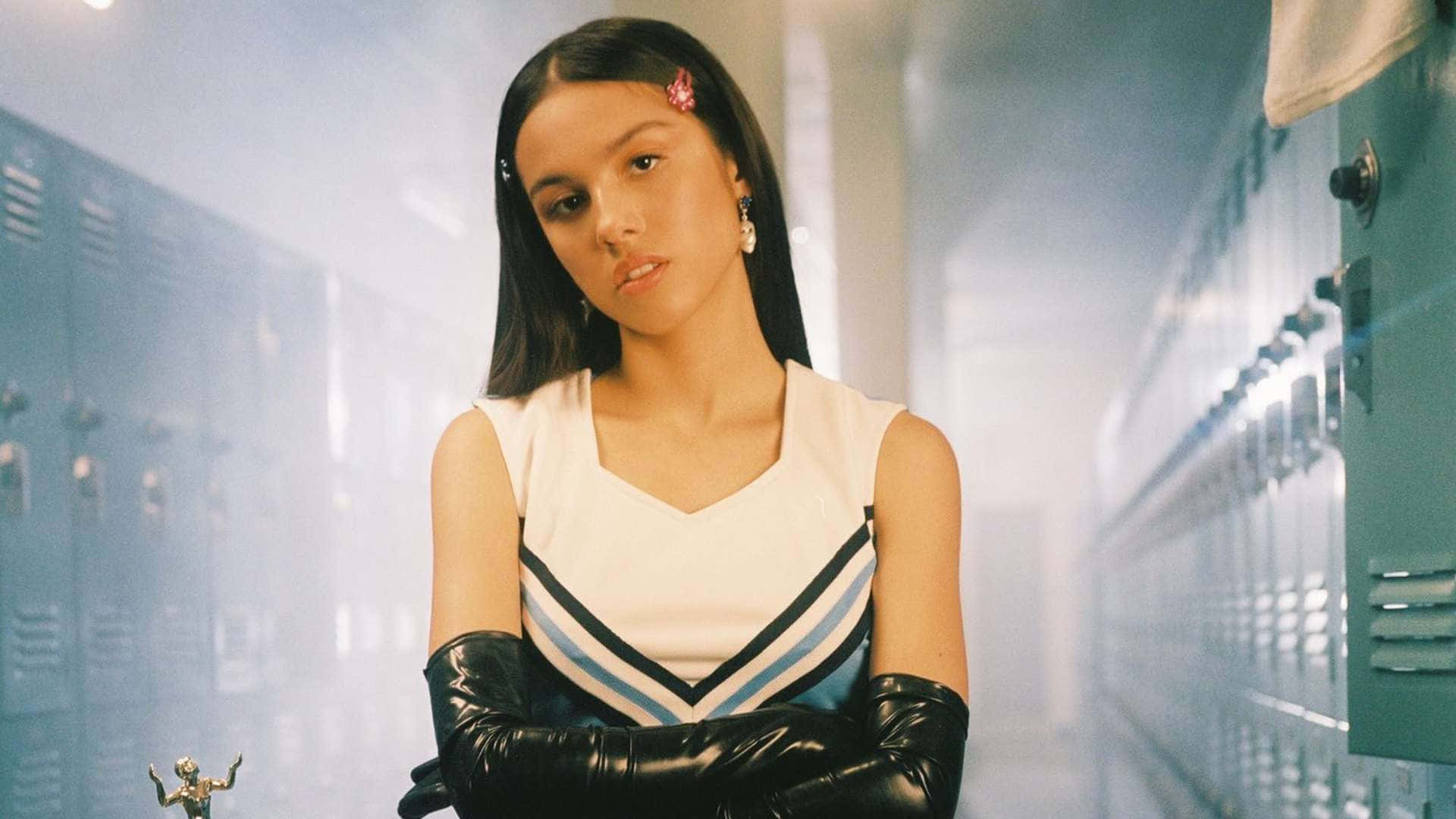 Olivia Rodrigo 桌布 壁紙 — 一位自信的啦啦隊員身穿白色制服和黑色手套，在霧氣瀰漫、兩旁有儲物櫃的學校走廊中交叉雙臂站立。