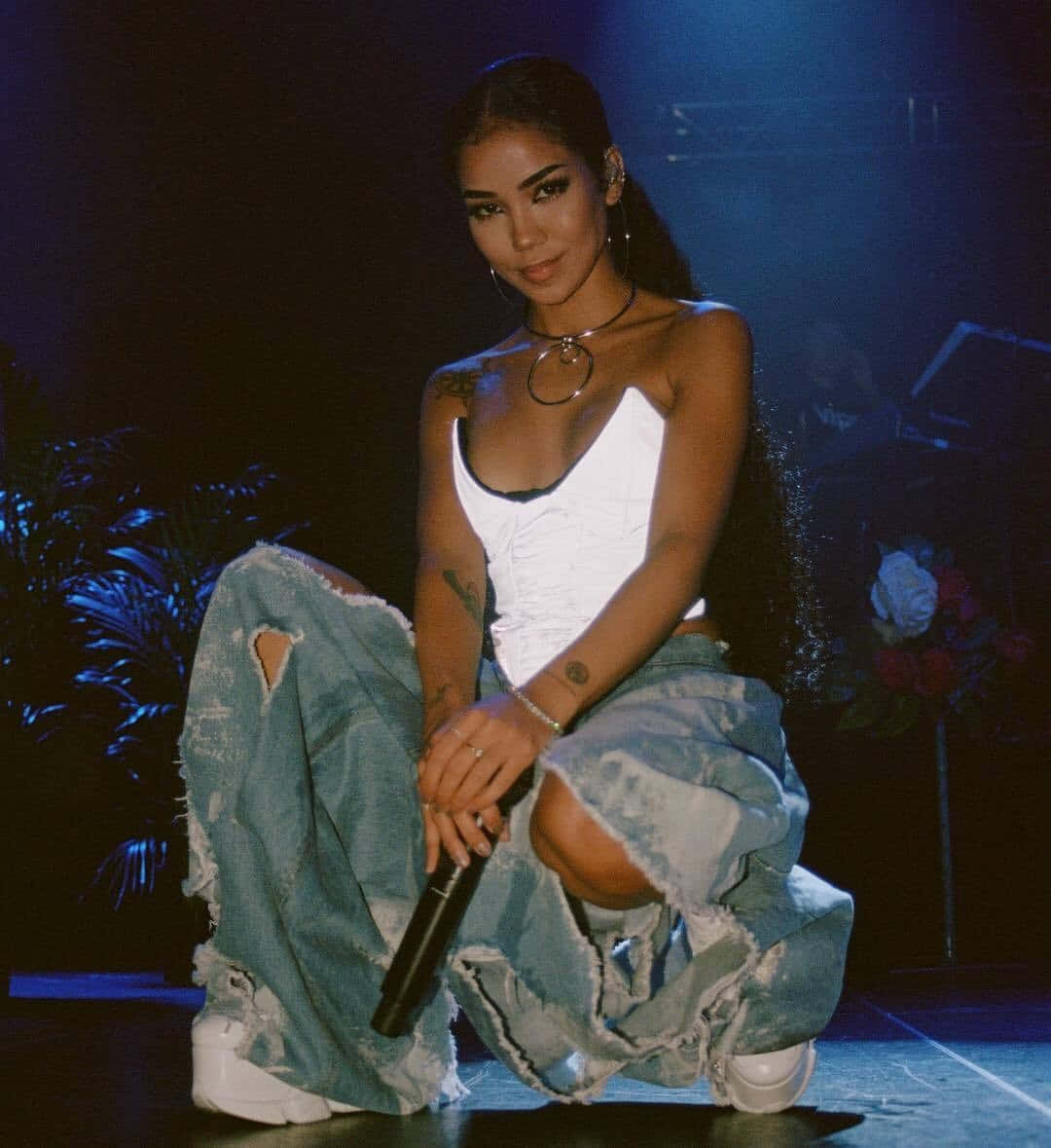 jhene aiko 桌布 壁紙 — 一位自信的女表演者穿著破洞牛仔褲和白色上衣，在藍色舞台燈光下跪在舞台上，手持麥克風。