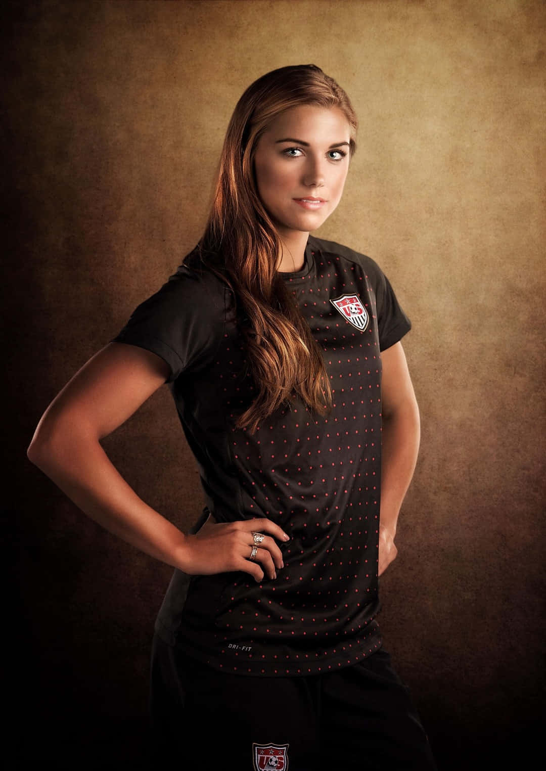 Alex Morgan 桌布 壁紙 — 一位自信的女足球員穿著黑色美國國家隊球衣，雙手叉腰，在帶有紋理的棕色背景前擺姿勢。