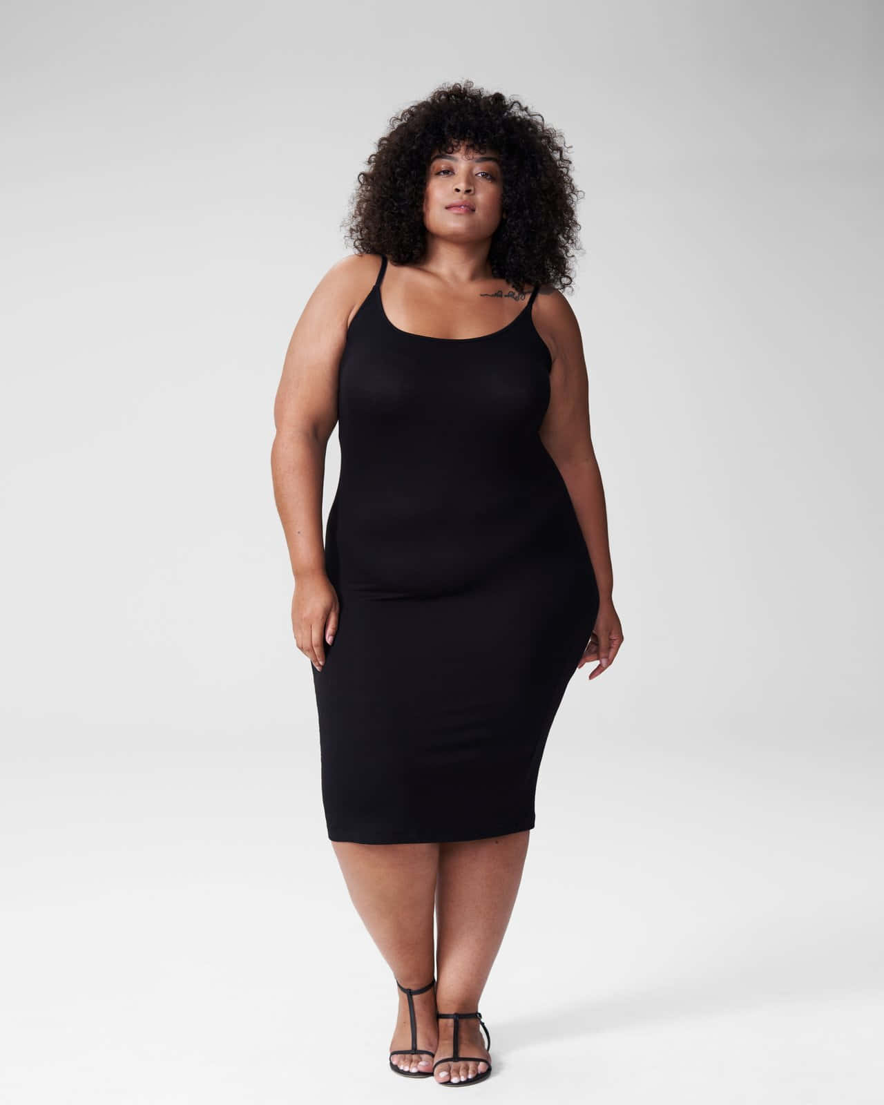 Minimalist Fashion 桌布 壁紙 — 一位自信的捲髮女性，身穿時尚黑色連衣裙和高跟鞋，背景為純白色。