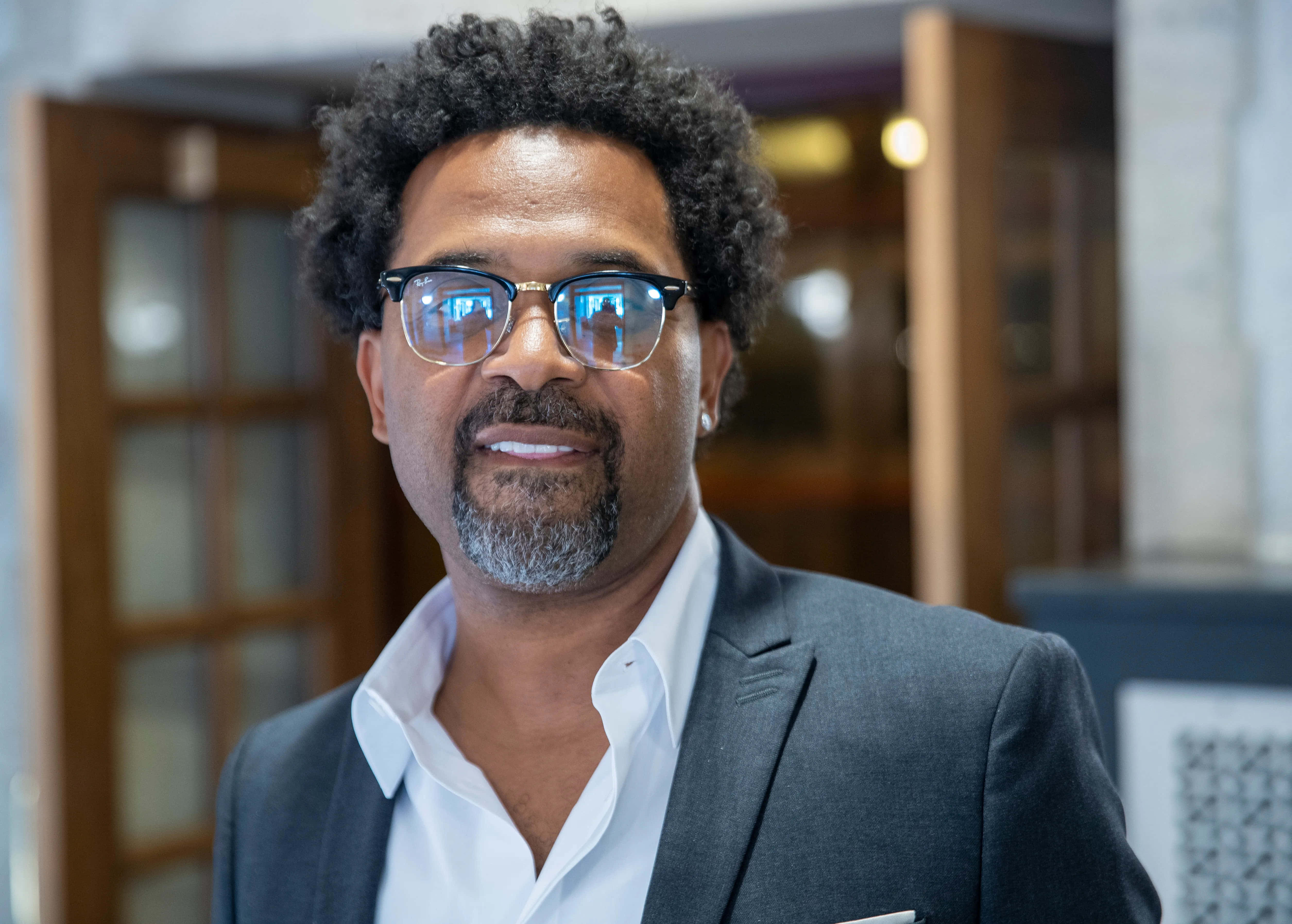 Mike Epps 桌布 壁紙 — 一位自信的男士，捲髮戴眼鏡，穿著時尚西裝，在現代室內環境中溫暖微笑。