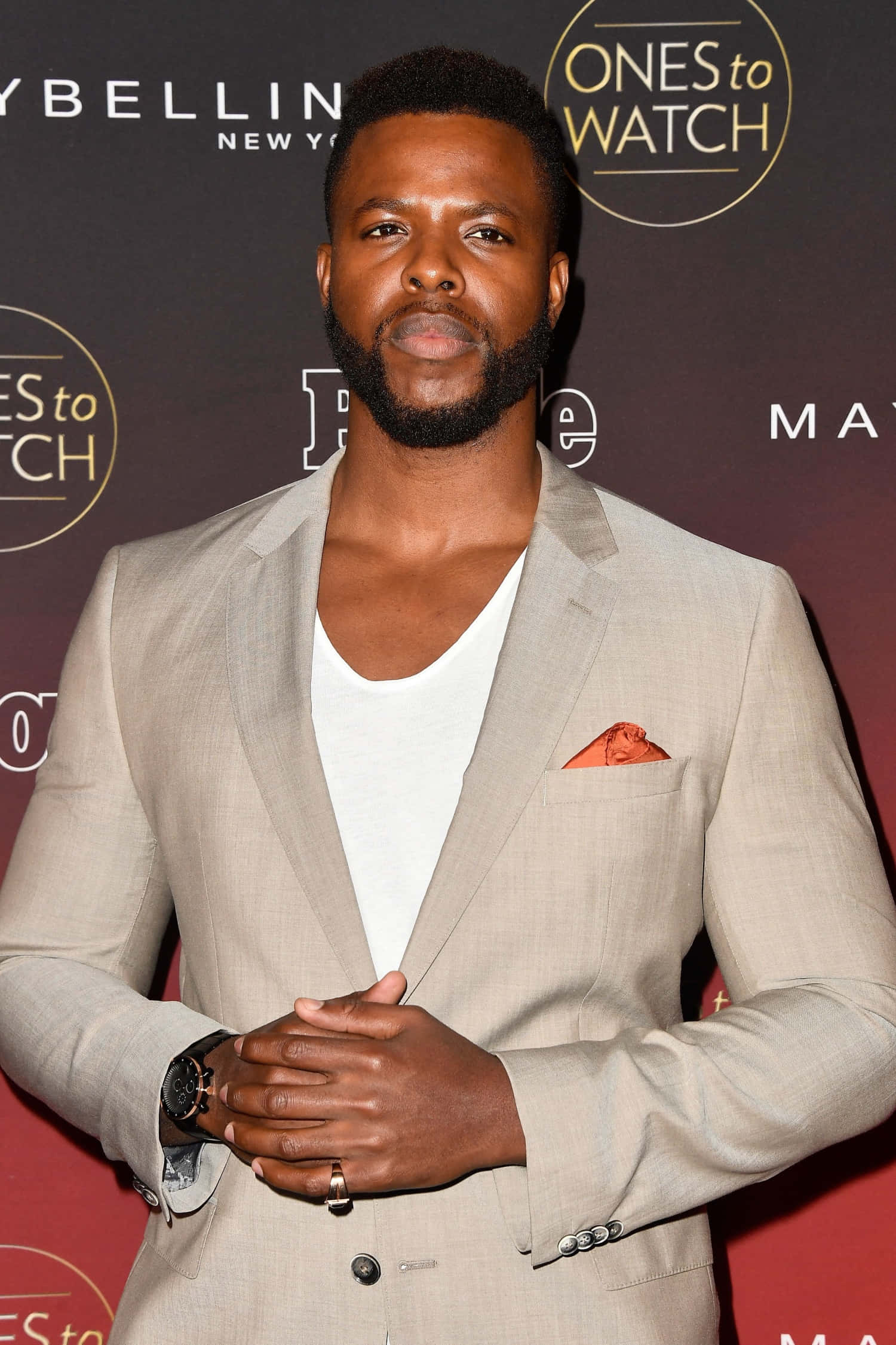 Winston Duke 桌布 壁紙 — 一位自信的男子身穿米色西裝和白色T恤，在有品牌背景的紅地毯活動中擺姿勢。