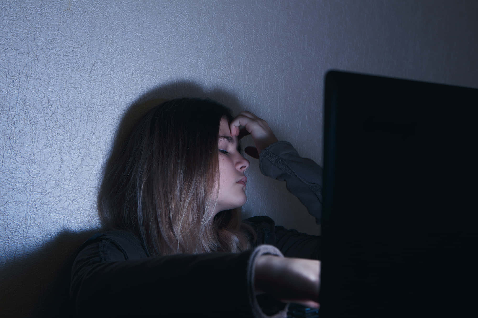 「一位與抑鬱奮鬥的年輕女子」 壁紙 — 一位年輕女性在昏暗光線中靠著牆，手放在額頭上，因盯著筆記型電腦螢幕而疲憊不堪。