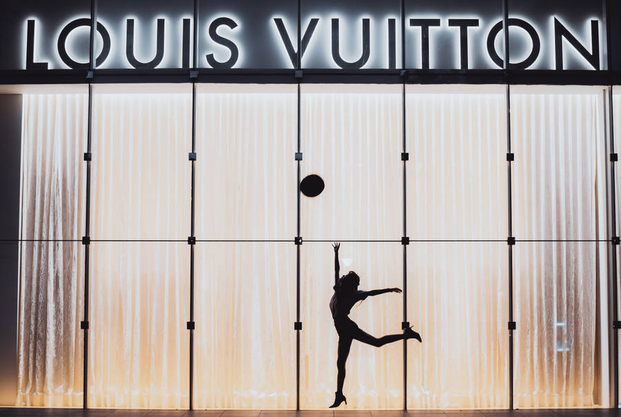 路易威登 桌布 壁紙 — 一位舞者的剪影在發光的 Louis Vuitton 店面（帶有垂直燈板）前優雅地跳躍。