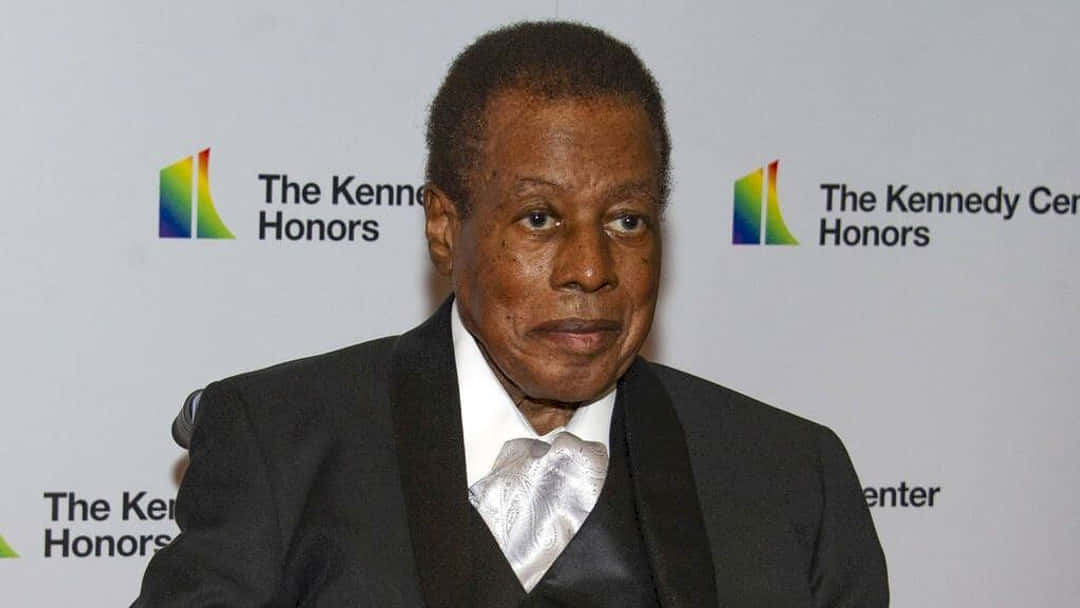 Wayne Shorter 桌布 壁紙 — 一位莊嚴的男子身穿黑色燕尾服，配銀色領帶，站在甘迺迪中心榮譽獎的背景前。
