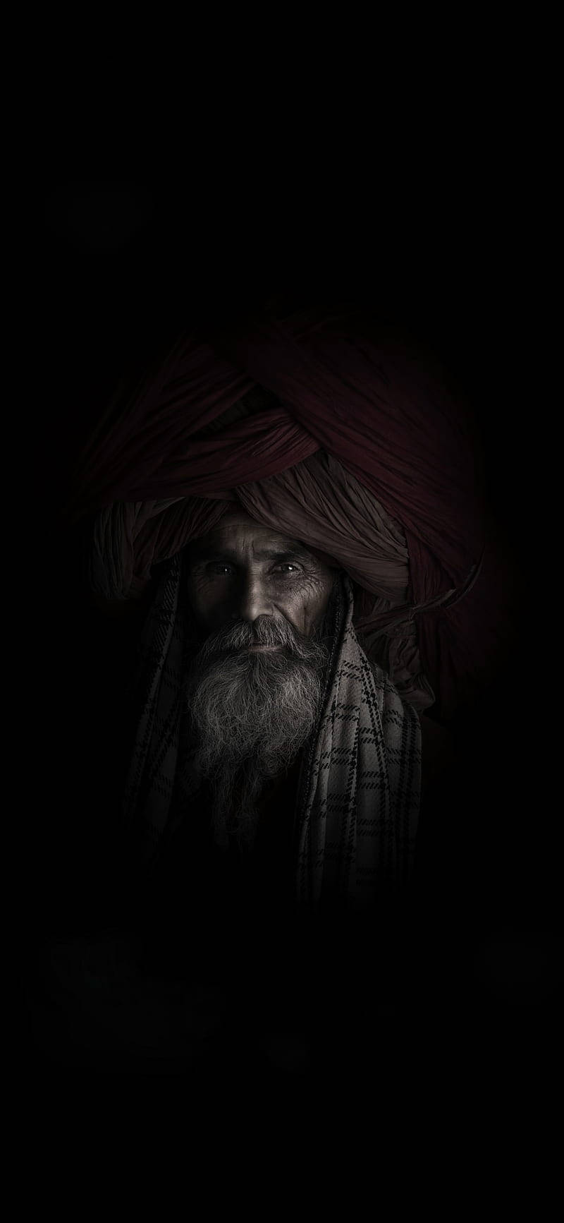 Aghori 桌布 壁紙 — 一位莊嚴的長者，留著長長的灰色鬍鬚，戴著紅色頭巾，在近乎黑暗的環境中凝視前方。