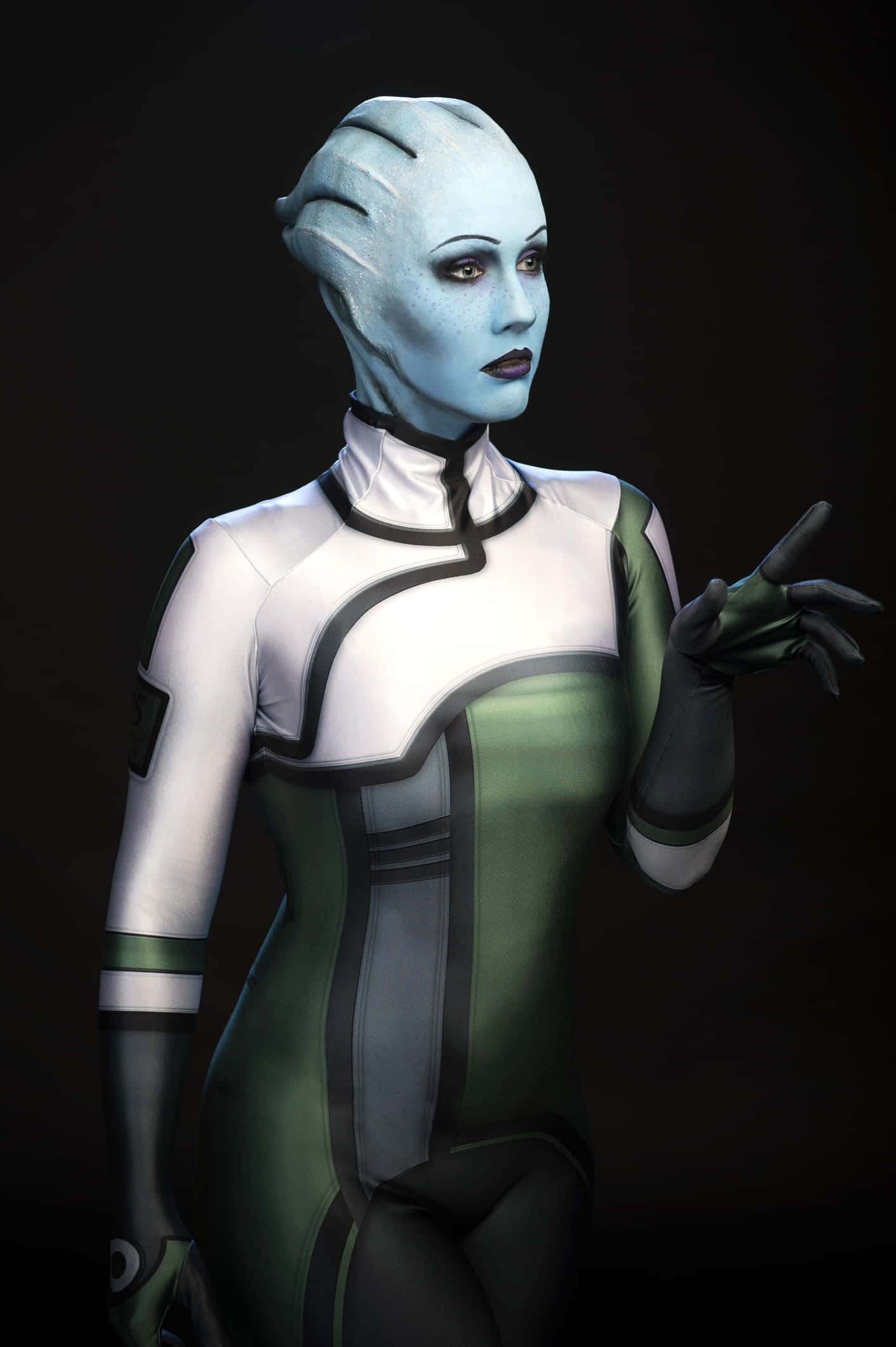 Liara T'soni 桌布 壁紙 — 一位藍皮膚的外星女性穿著白色和綠色的未來風格套裝，在深色背景前做手勢。