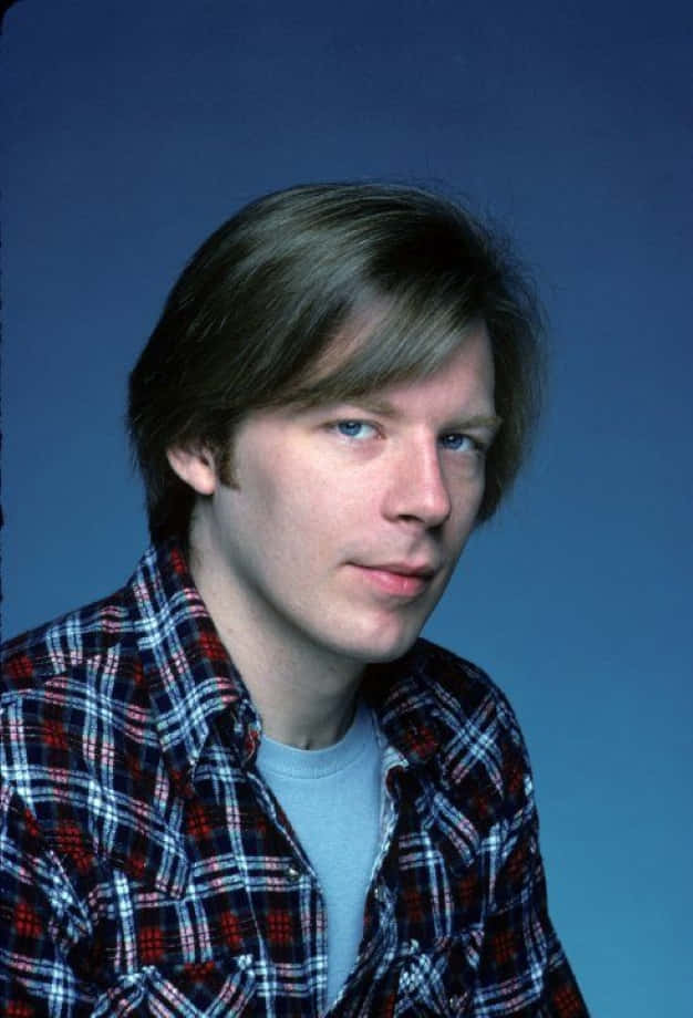 Michael Mckean 桌布 壁紙 — 一位藍眼睛、棕色頭髮的年輕男子穿著紅藍白格仔襯衫，背景是純藍色攝影棚。