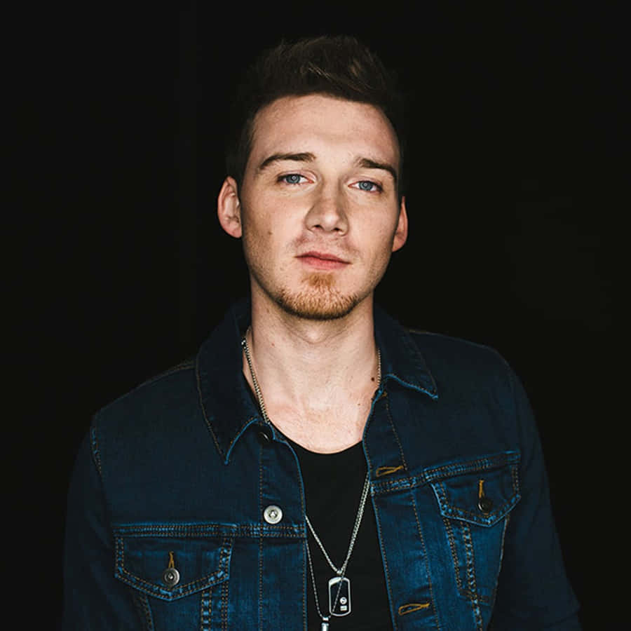 Morgan Wallen 桌布 壁紙 — 一位藍眼睛、鬍渣的年輕男子，在深色背景前穿著黑色襯衫外搭牛仔外套。