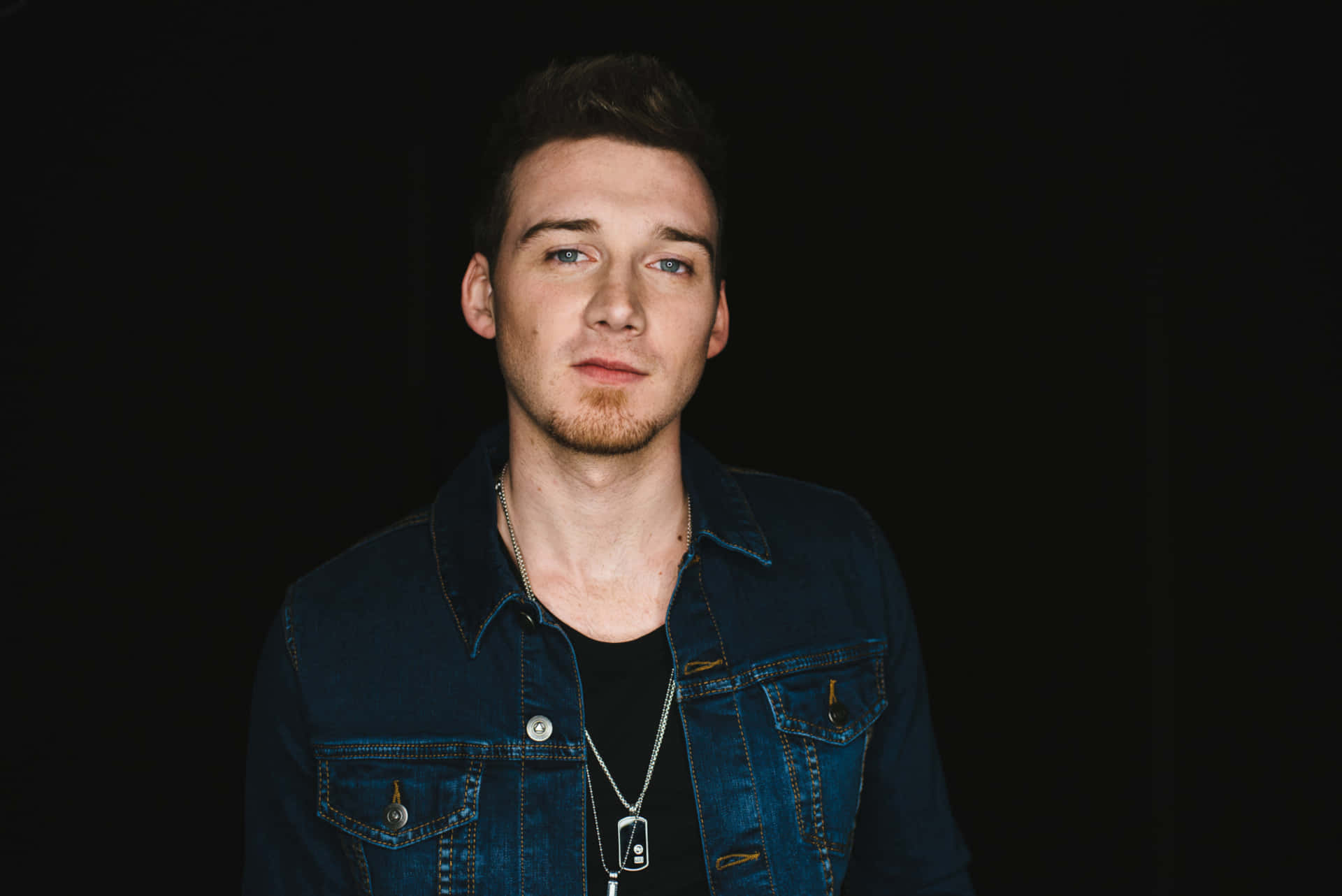 Morgan Wallen 桌布 壁紙 — 一位藍眼睛、鬍渣的年輕男子，穿著牛仔外套，背景為黑色。