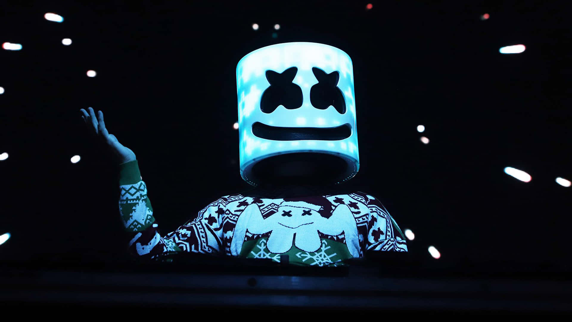 Marshmello 桌布 壁紙 — 一位表演者戴著發光的藍色棉花糖頭盔和節日毛衣，在一個有漂浮燈光的黑暗演唱會舞台上揮手。