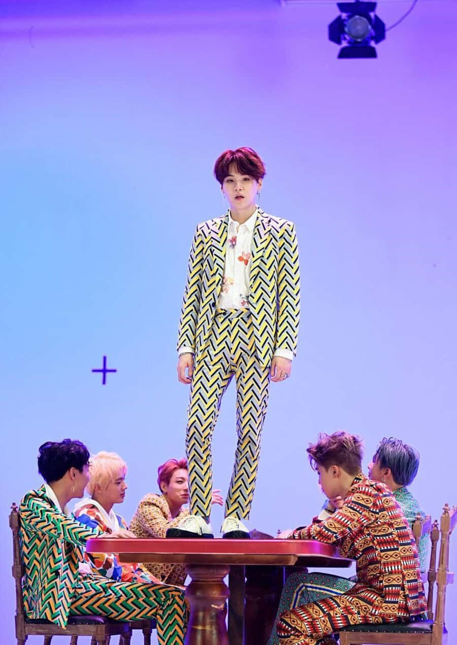 Bts Mv 桌布 壁紙 — 一位表演者穿著大膽的黃色鋸齒紋西裝站在桌子上，另外五位穿著彩色圖案西裝的人坐在周圍，舞台燈光為紫色。