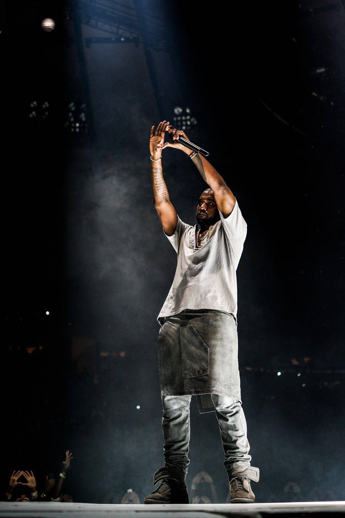Kanye West All Grey Shirt 壁紙 — 一位表演者站在舞台中央，高舉雙臂握著麥克風，在戲劇性的聚光燈下，煙霧在他周圍旋轉。