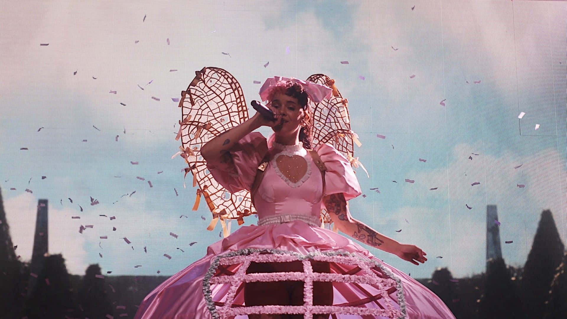 Melanie Martinez 桌布 壁紙 — 一位表演者身穿寬大的粉紅色長袍，帶有編織翅膀，對著麥克風唱歌，五彩紙屑在陰天飄落。