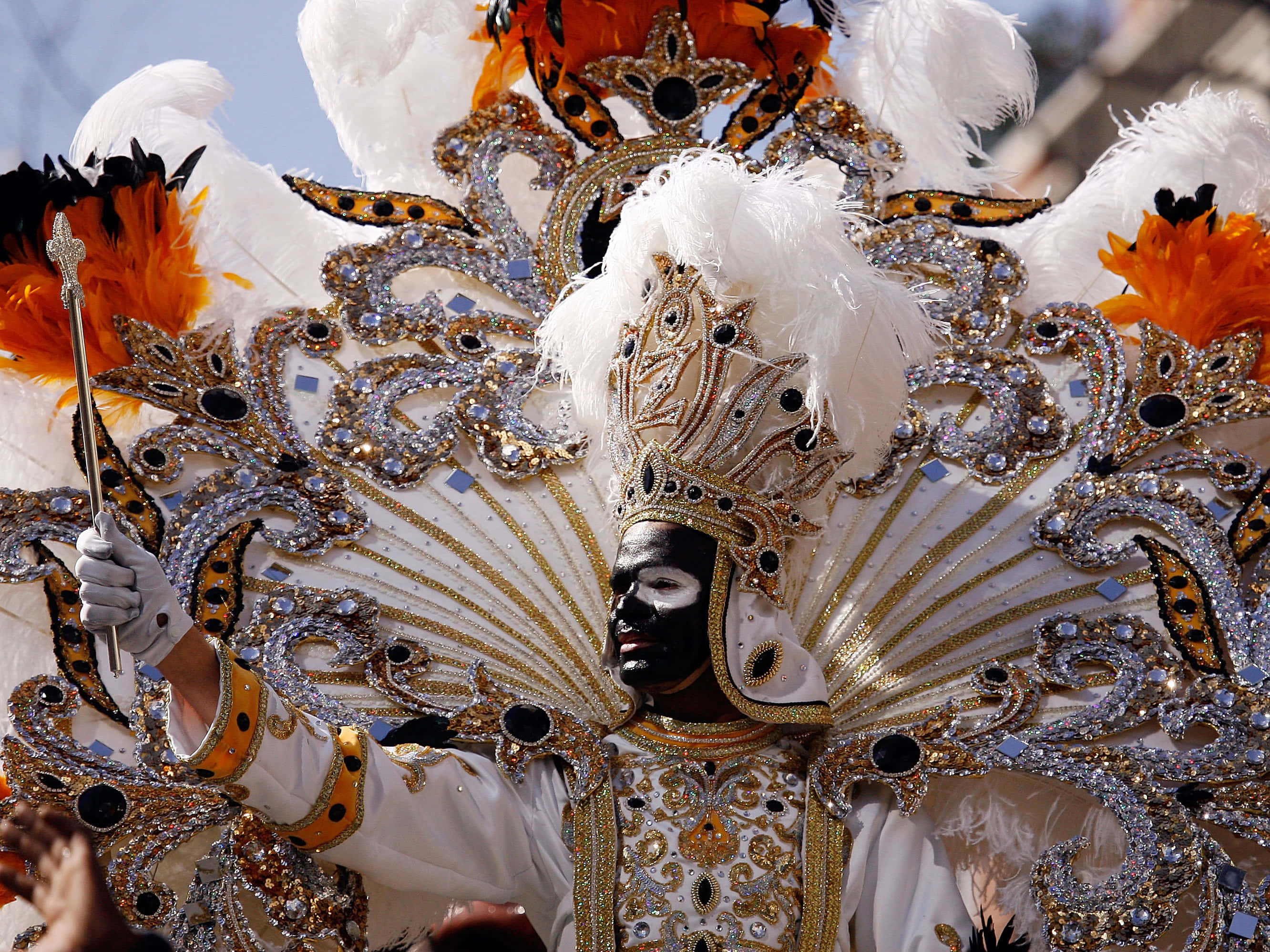 Mardi Gras 桌布 壁紙 — 一位表演者身穿華麗的白金色和橙色羽毛服飾，臉上塗著黑色顏料，在充滿活力的嘉年華遊行中手持權杖。