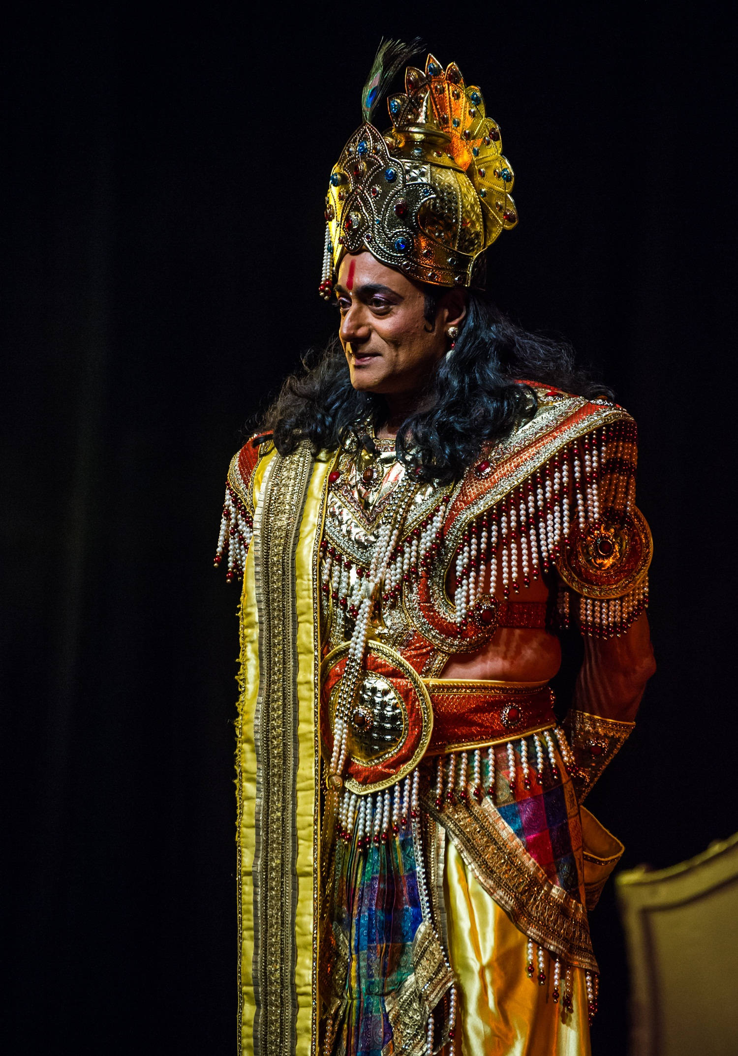 krishna iPhone 桌布 壁紙 — 一位表演者身穿華麗的金色和紅色傳統服飾，頭戴珠寶頭飾，在黑暗背景下微笑。