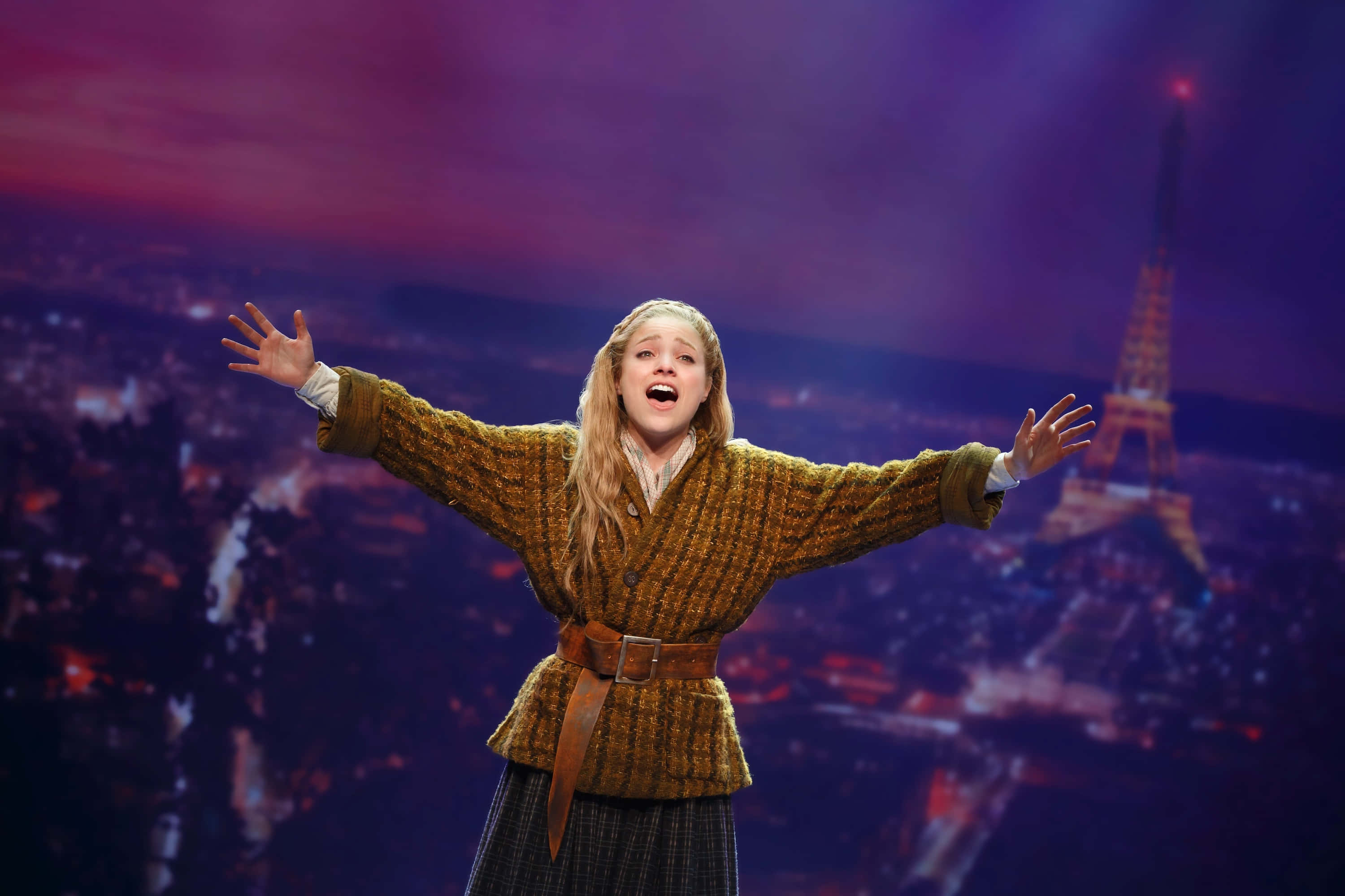 Anastasia 桌布 壁紙 — 一位表演者身穿質感棕色外套，在充滿活力的紫色暮色背景下張開雙臂，背景是艾菲爾鐵塔。