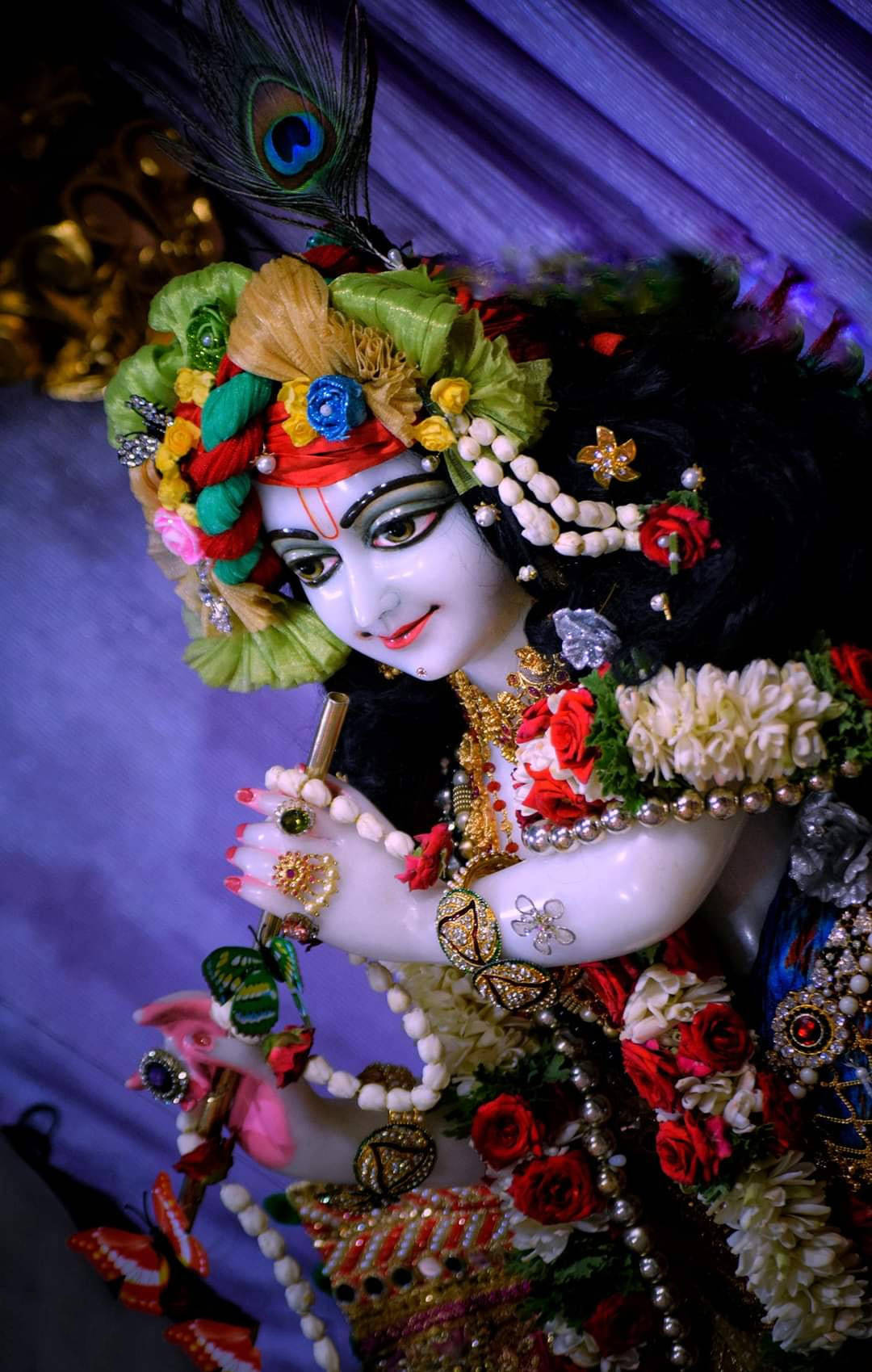 krishna god 桌布 壁紙 — 一位裝飾華麗的神祇，頭戴孔雀羽毛頭飾，身披花環，在深紫色垂簾背景下手持長笛。