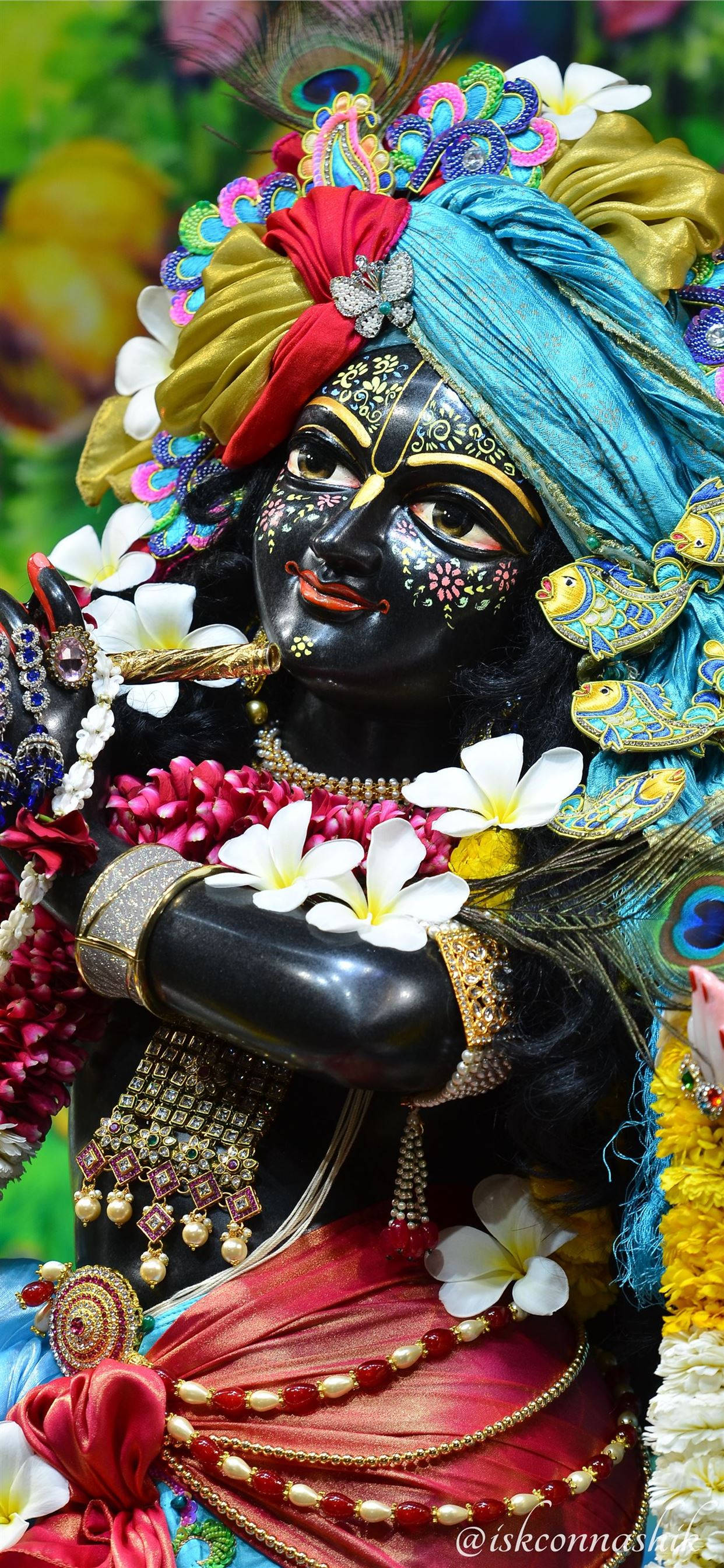 krishna god 桌布 壁紙 — 一位裝飾華麗的黑皮膚神祇，佩戴精緻的金飾和色彩繽紛的花環，背景是茂密的綠色。