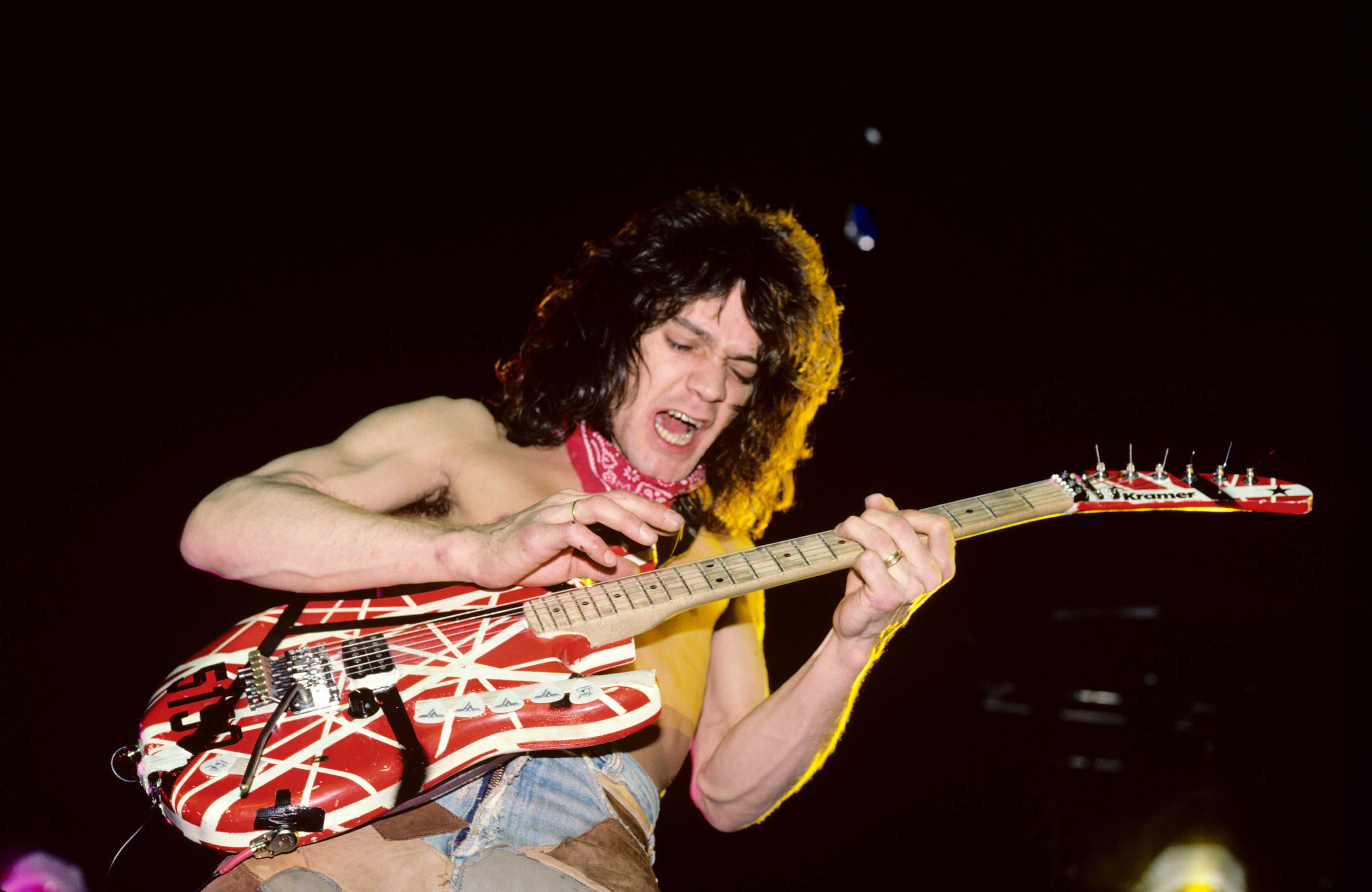 Eddie Van Halen Frankenstrat 壁紙 — 一位赤裸上身的結他手，捲髮，在舞台燈光下熱情地彈奏紅白電結他。