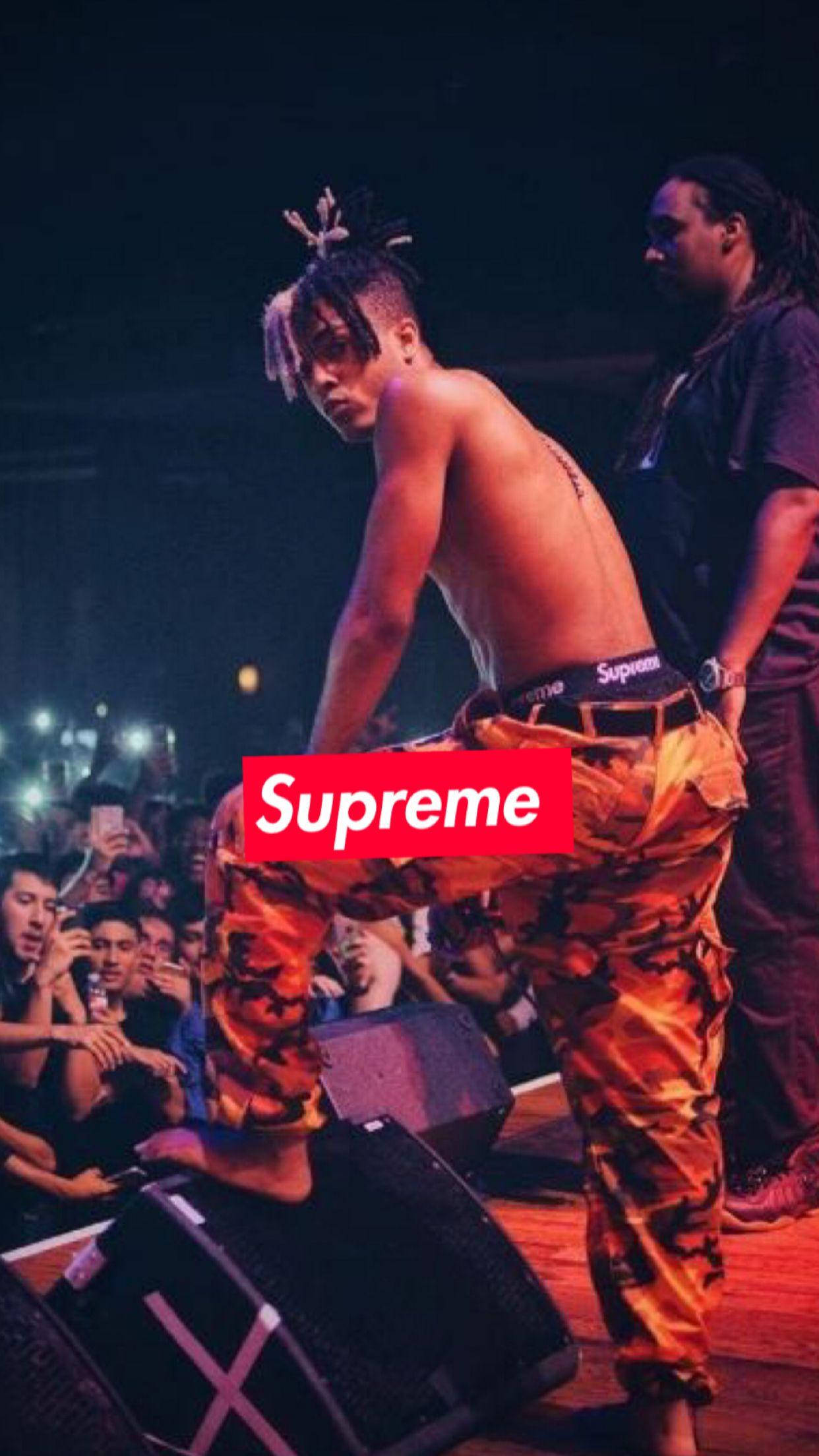 XXXTENTACION 桌布 壁紙 — 一位赤裸上身的表演者穿著橙色迷彩褲和Supreme皮帶，在昏暗的舞台上掌控全場，面對投入的觀眾。